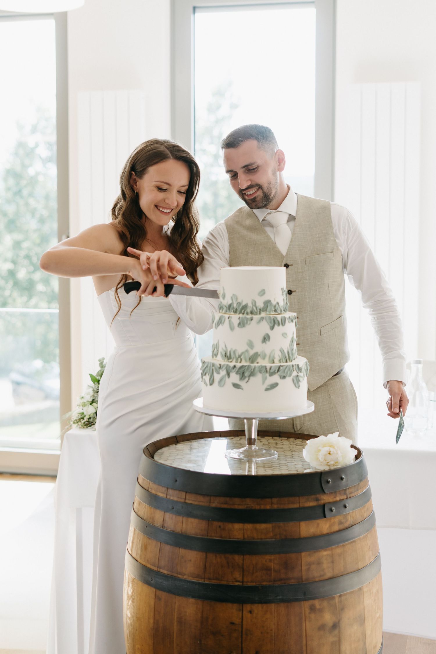 Couple cutting their elegant three-tier white wedding cake displayed on rustic wine barrel at bright indoor venue.