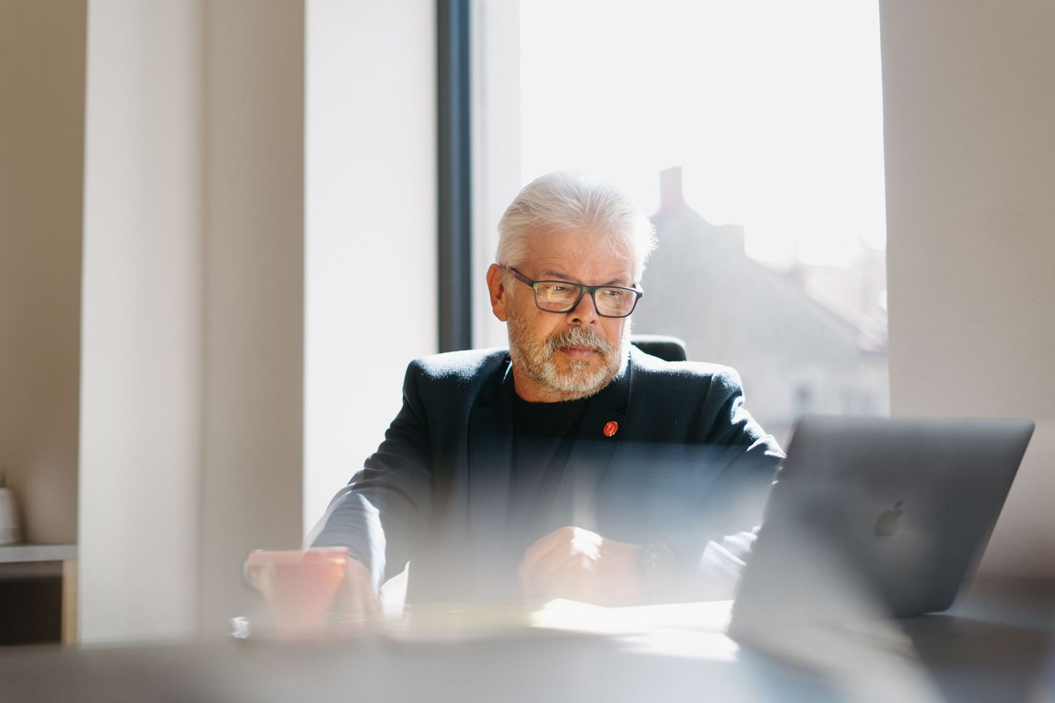 Individual wearing glasses works at a laptop computer in an office with natural window lighting casting a soft glow.