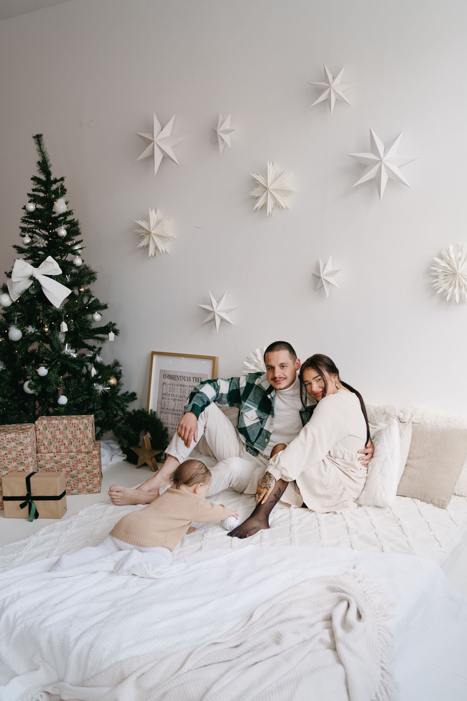 Cozy Christmas bedroom scene with white bedding, decorated tree and paper star ornaments on the wall.