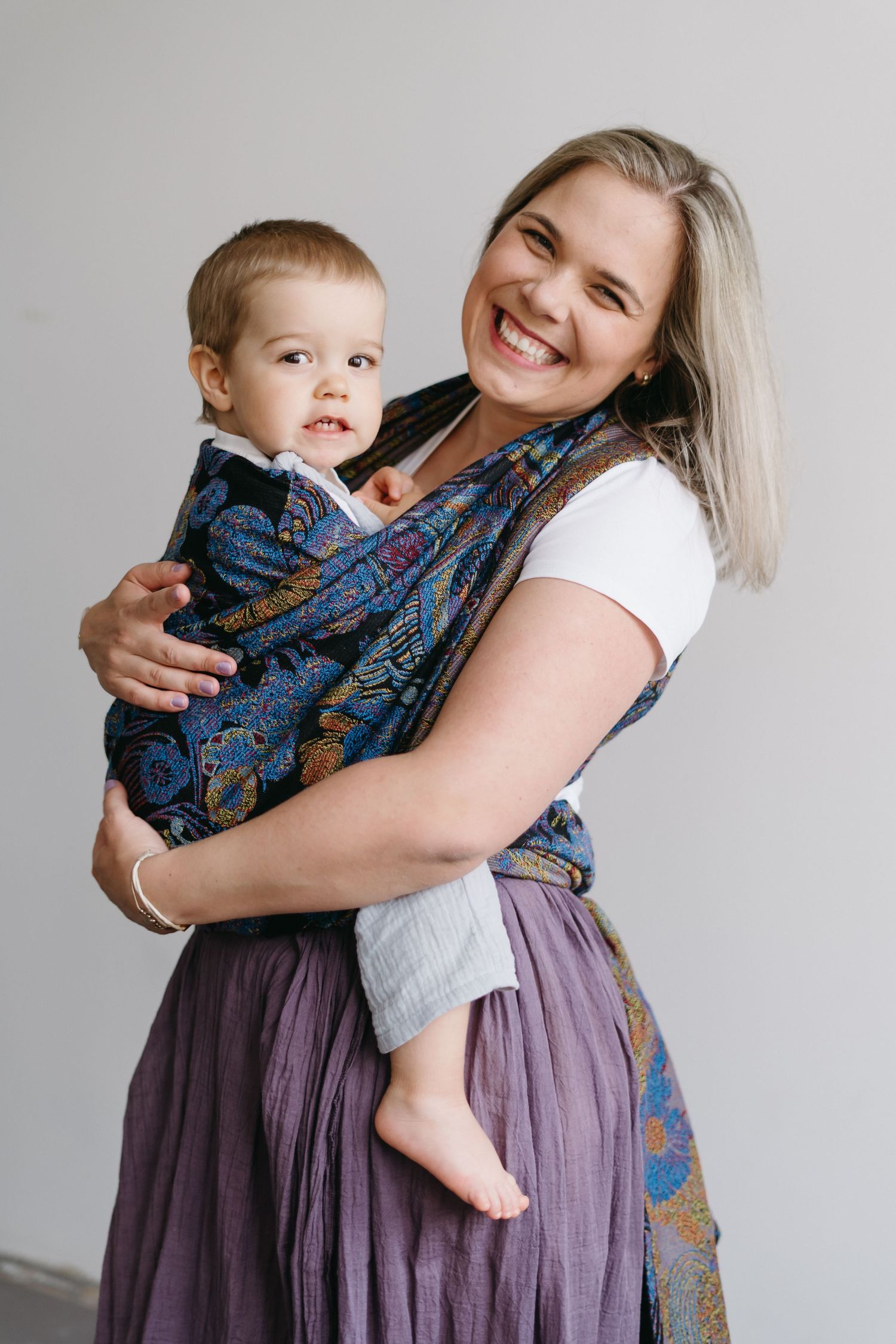 Smiling woman wearing purple skirt carries toddler in patterned blue baby carrier against white wall.