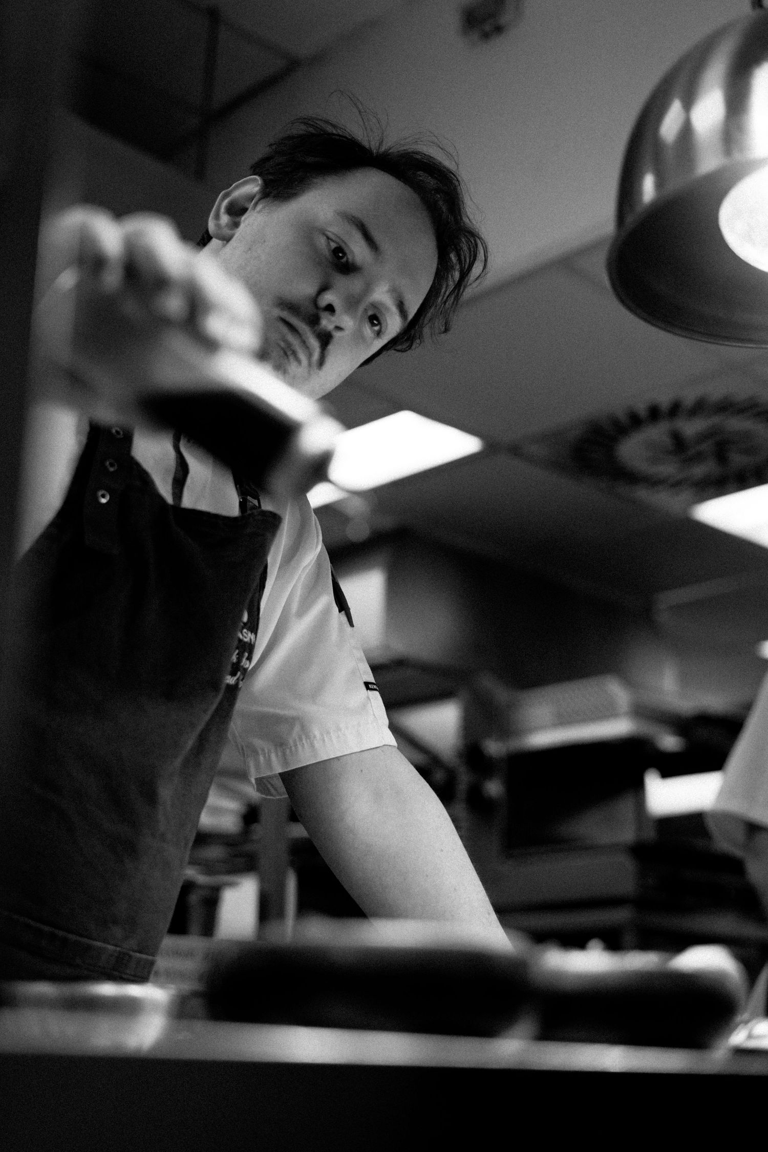 Black and white photo of a worker focusing intently on food preparation in a restaurant kitchen under overhead lighting.