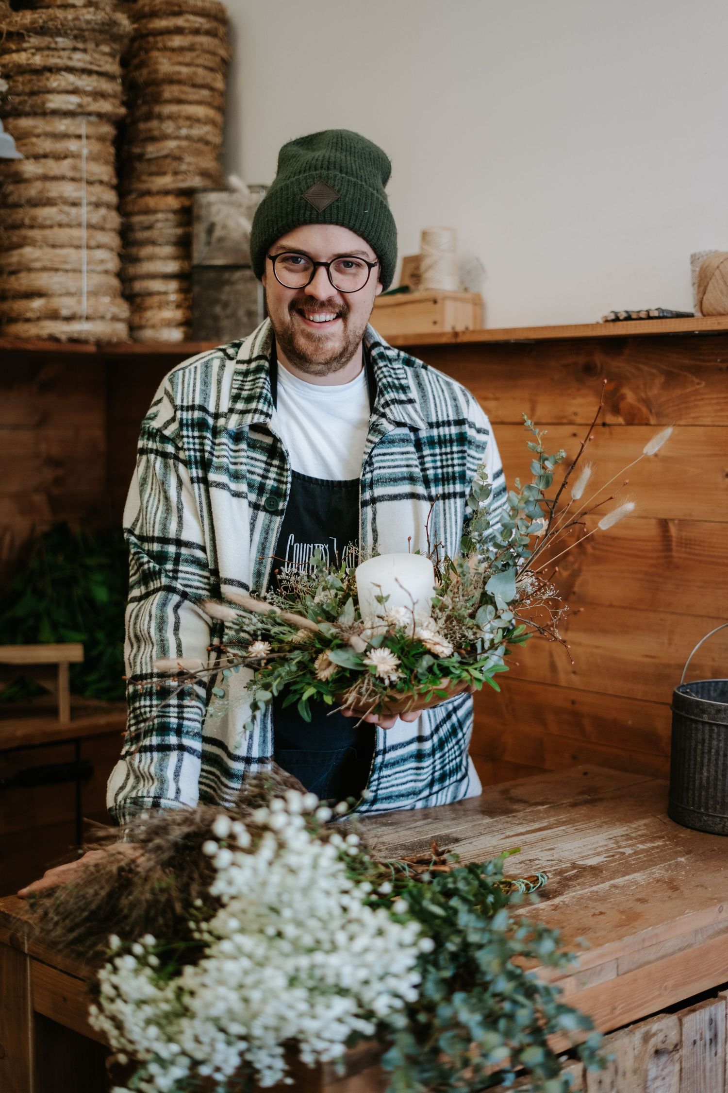 Smiling bearded man in glasses, beanie and plaid shirt holds a dried flower wreath with a candle at a rustic wooden florist workshop.