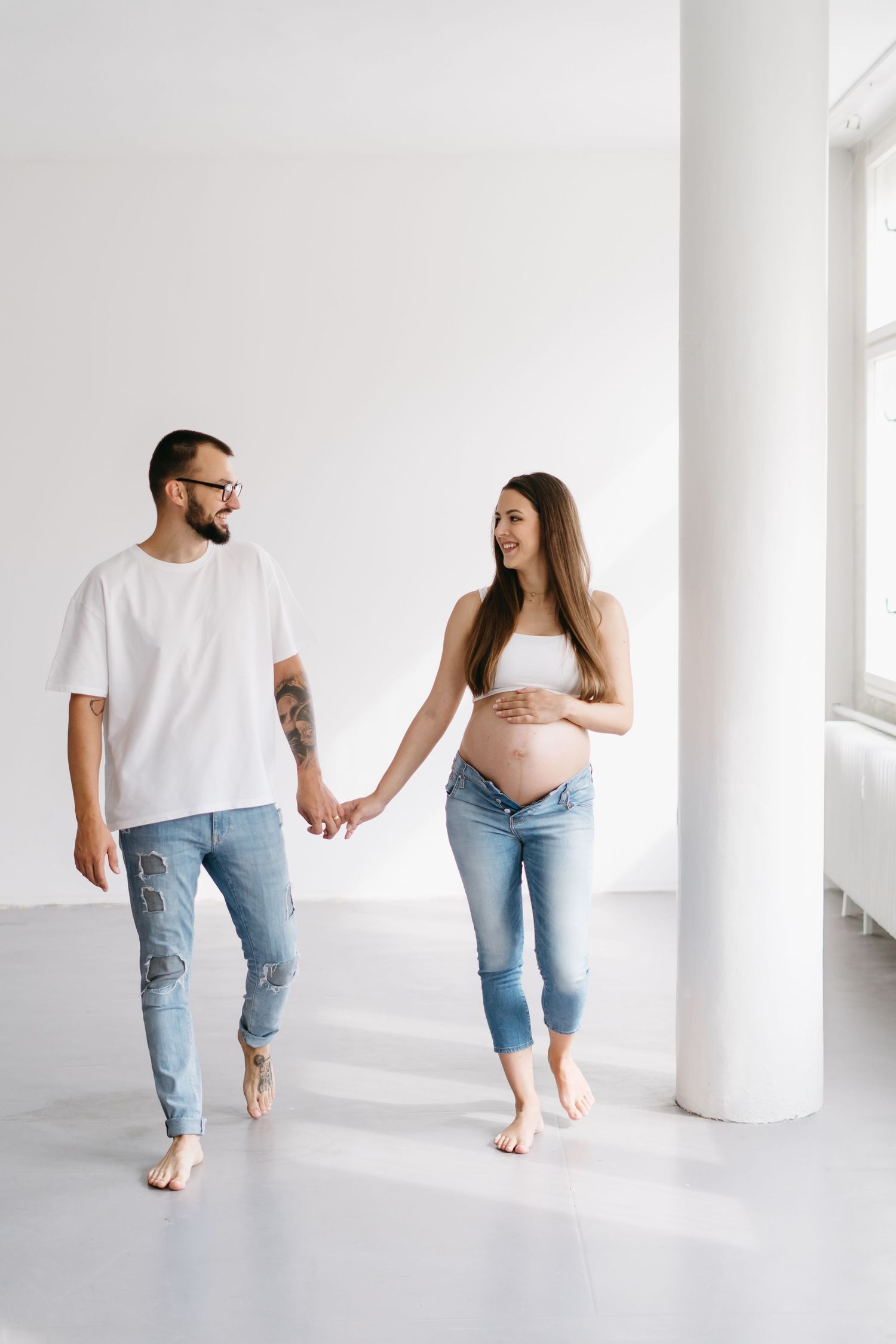 Couple walking barefoot together against white wall in casual denim outfits.