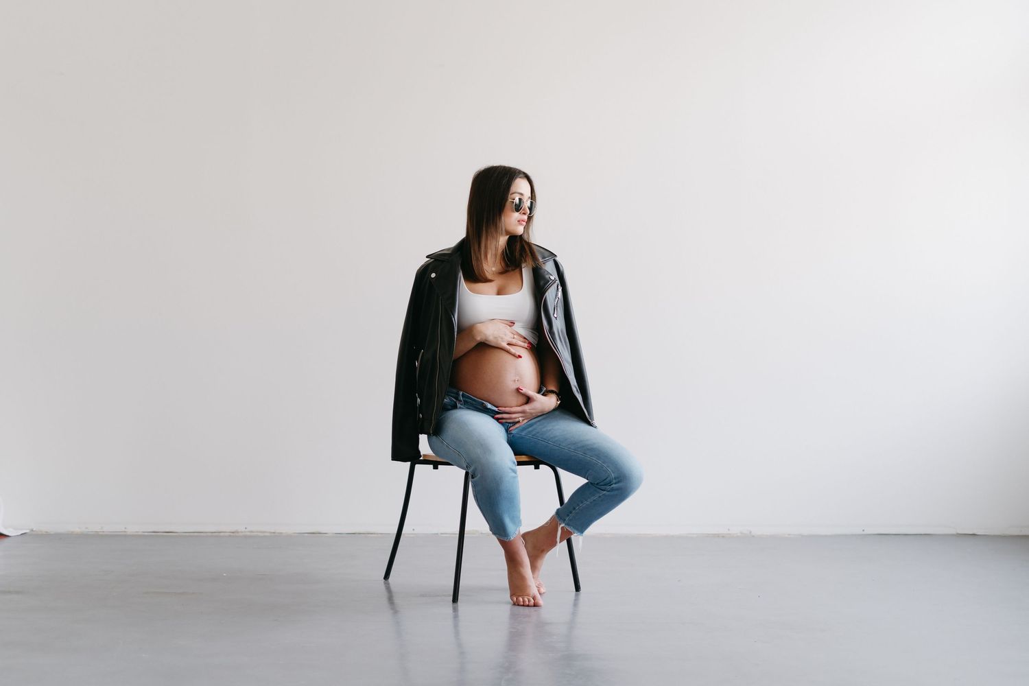 Modern maternity portrait with woman seated on chair in minimalist studio setting.
