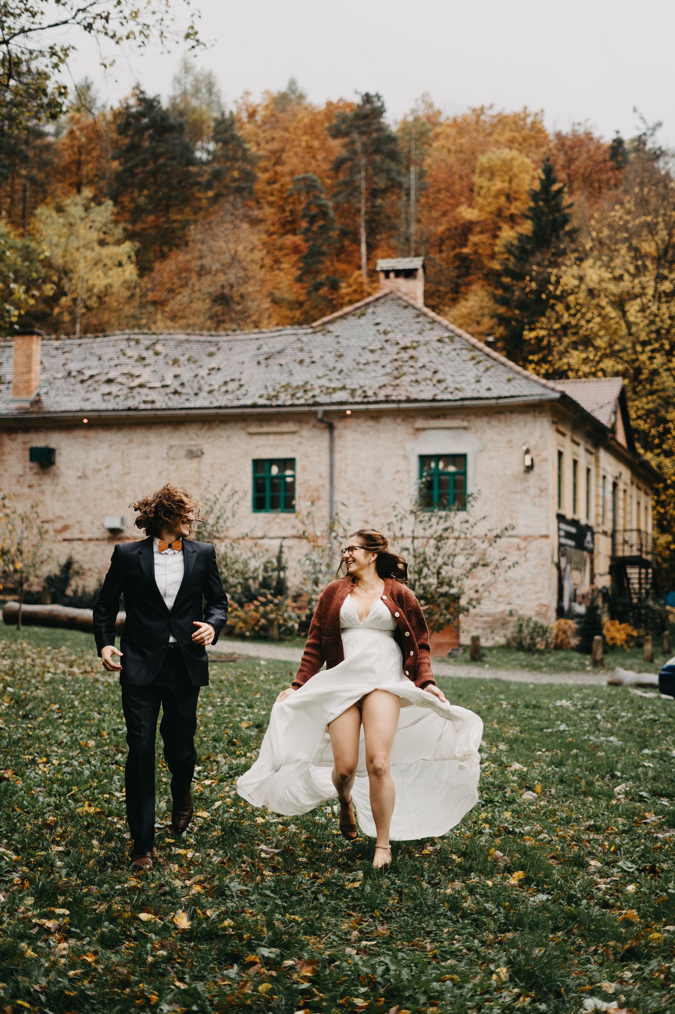 A romantic autumn wedding scene with a couple running joyfully in front of a rustic stone building surrounded by fall foliage.