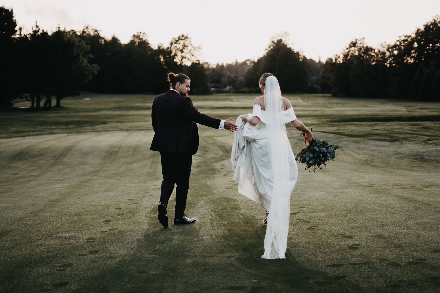 A couple in wedding attire holds hands while walking on a grassy field at sunset.