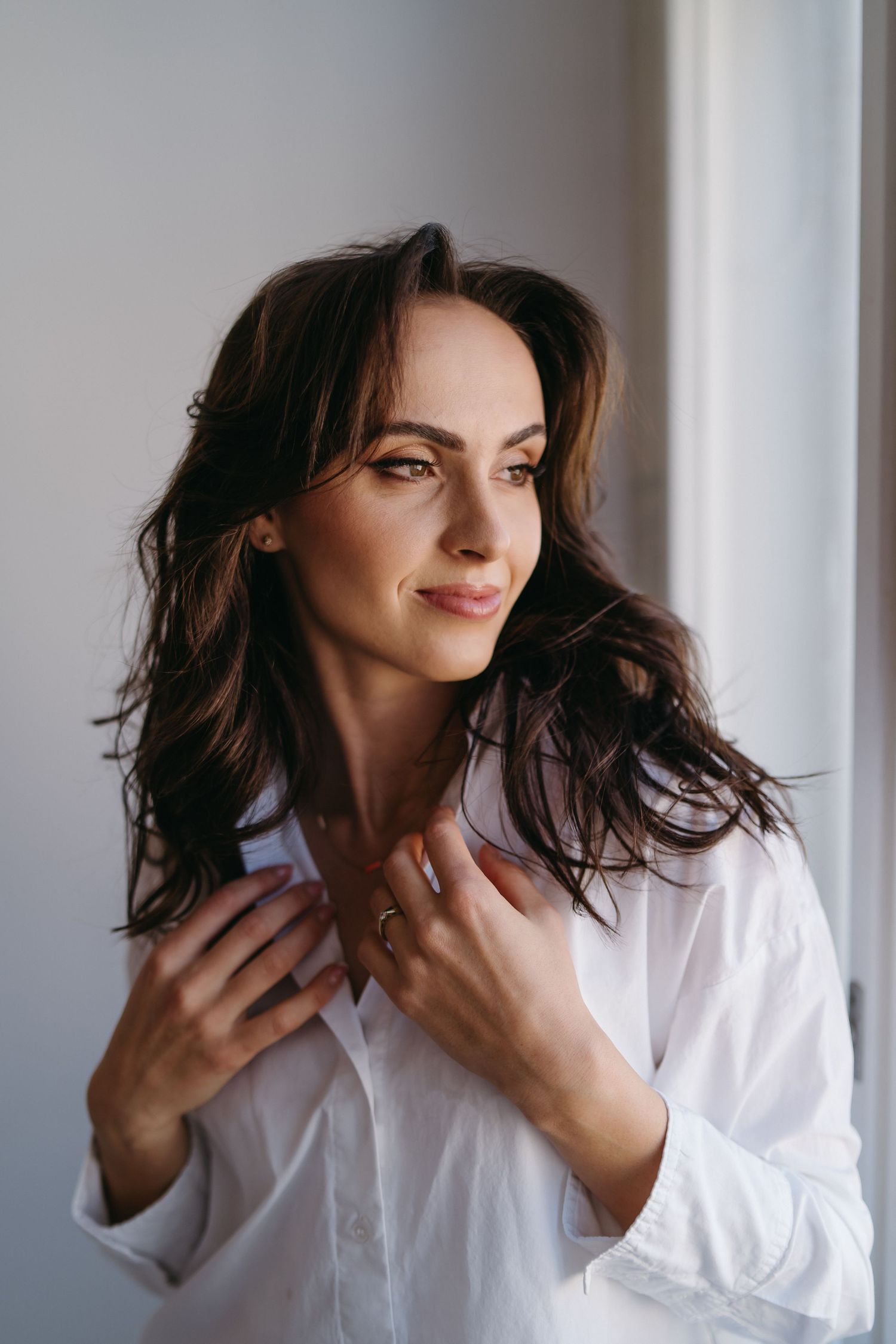 A brunette woman in a white shirt holds her collar while gazing sideways near a bright window.