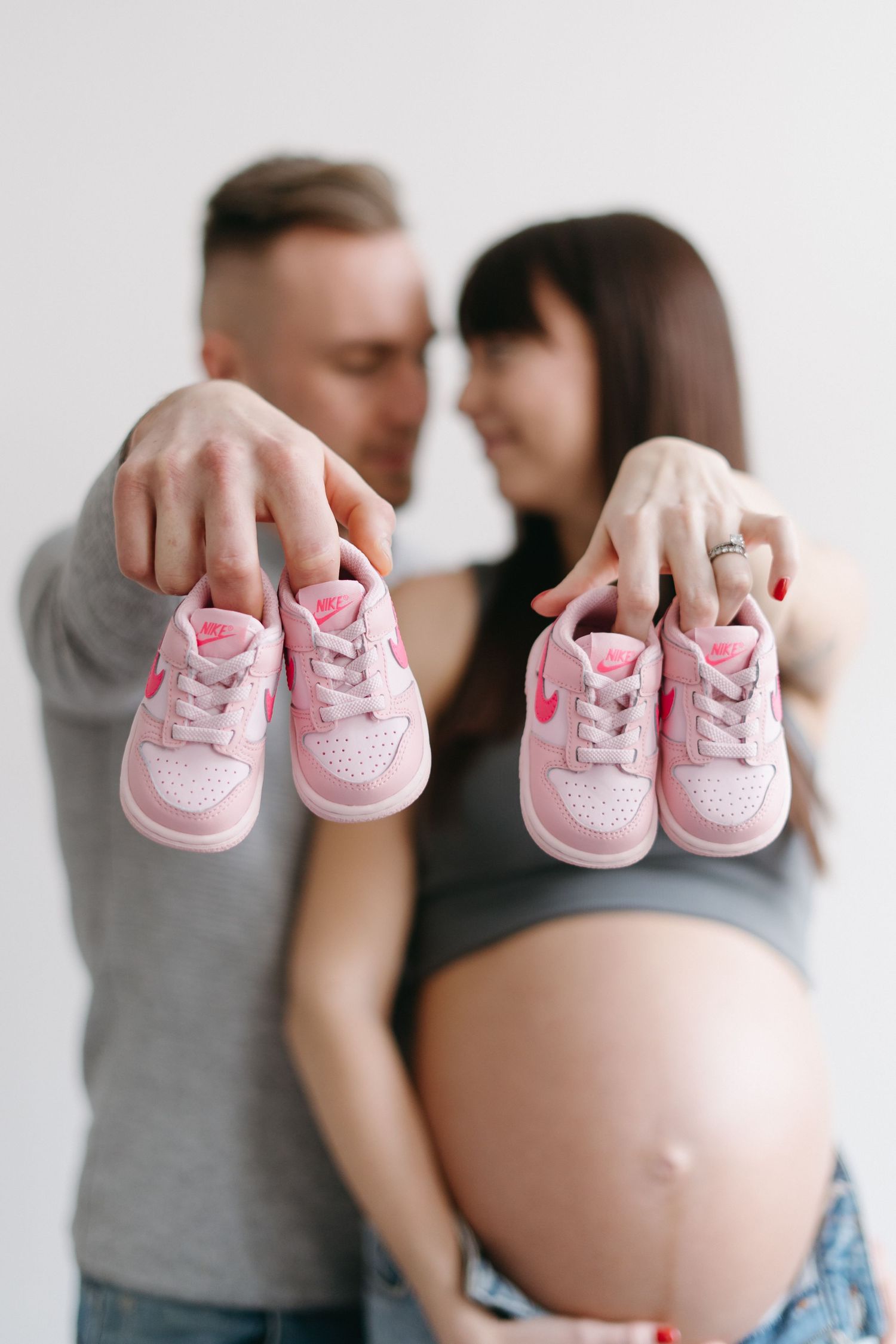 Couple holding tiny pink sneakers in front of pregnant belly against minimal white background.