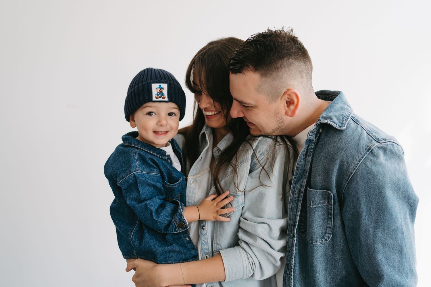 Happy young family with toddler boy in denim jacket and beanie smiling together against white background.