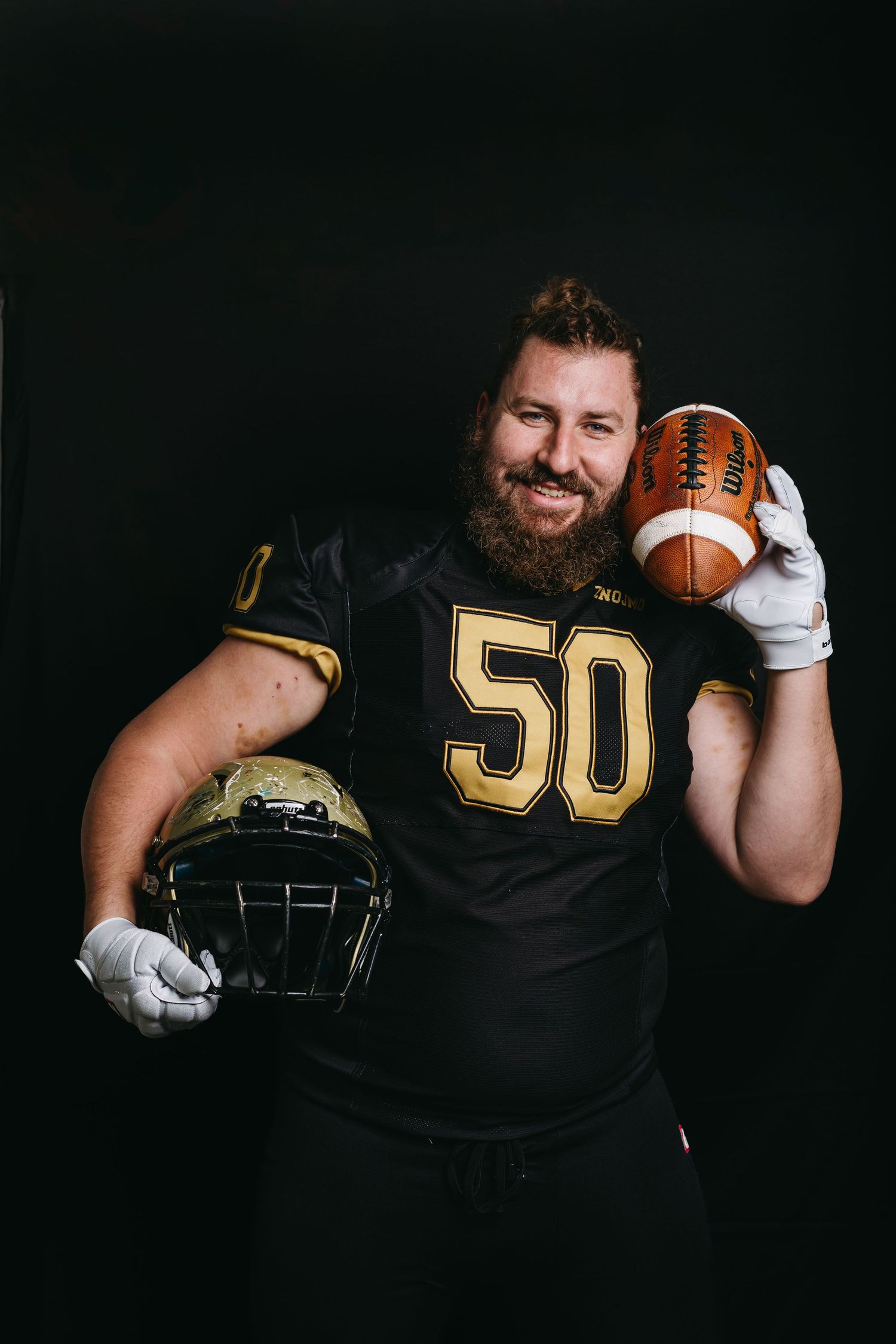 Bearded football player wearing black jersey number 50 holds football and helmet against dark background.