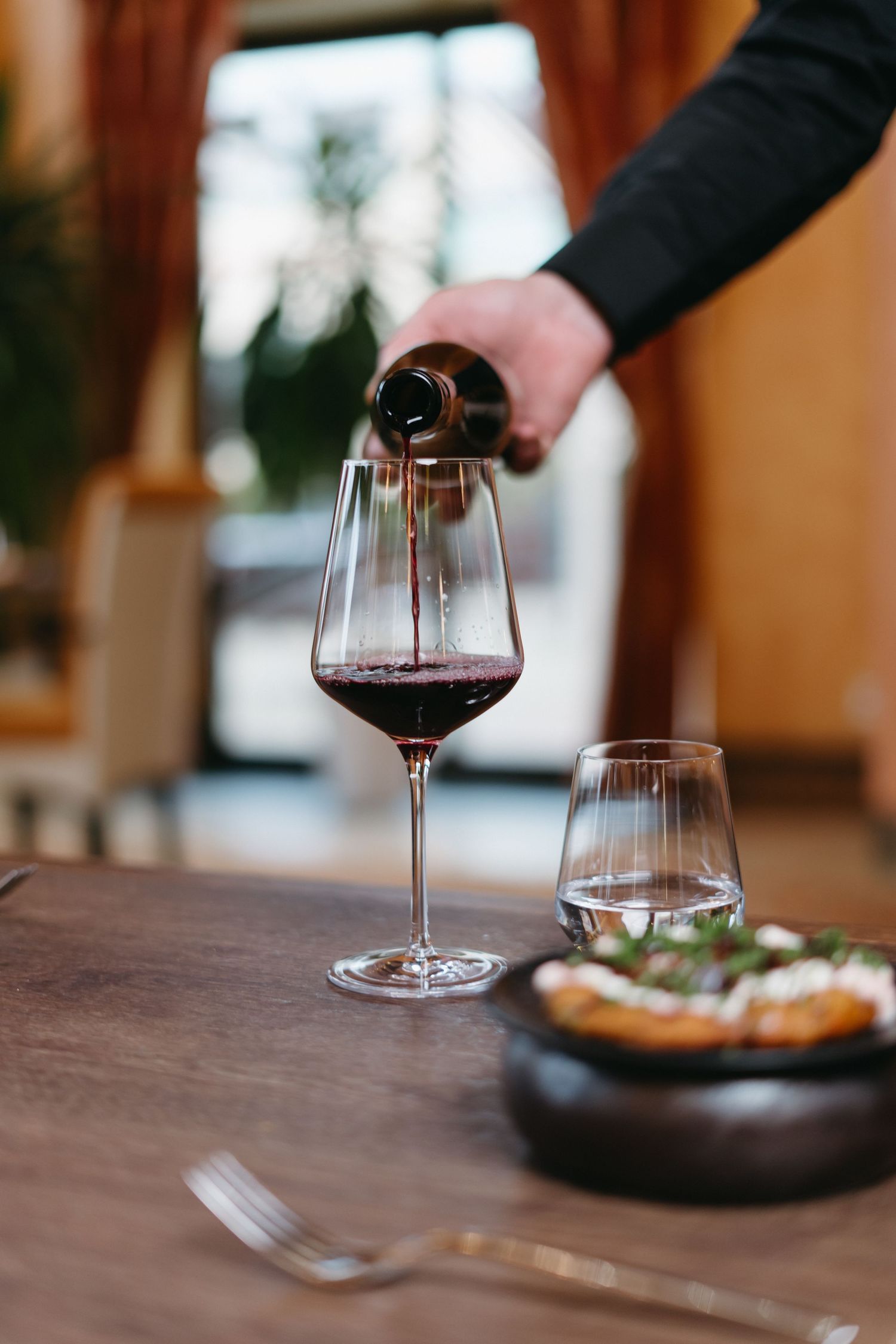 Hand pouring red wine into an elegant stemmed glass on a wooden dining table with food in the background.