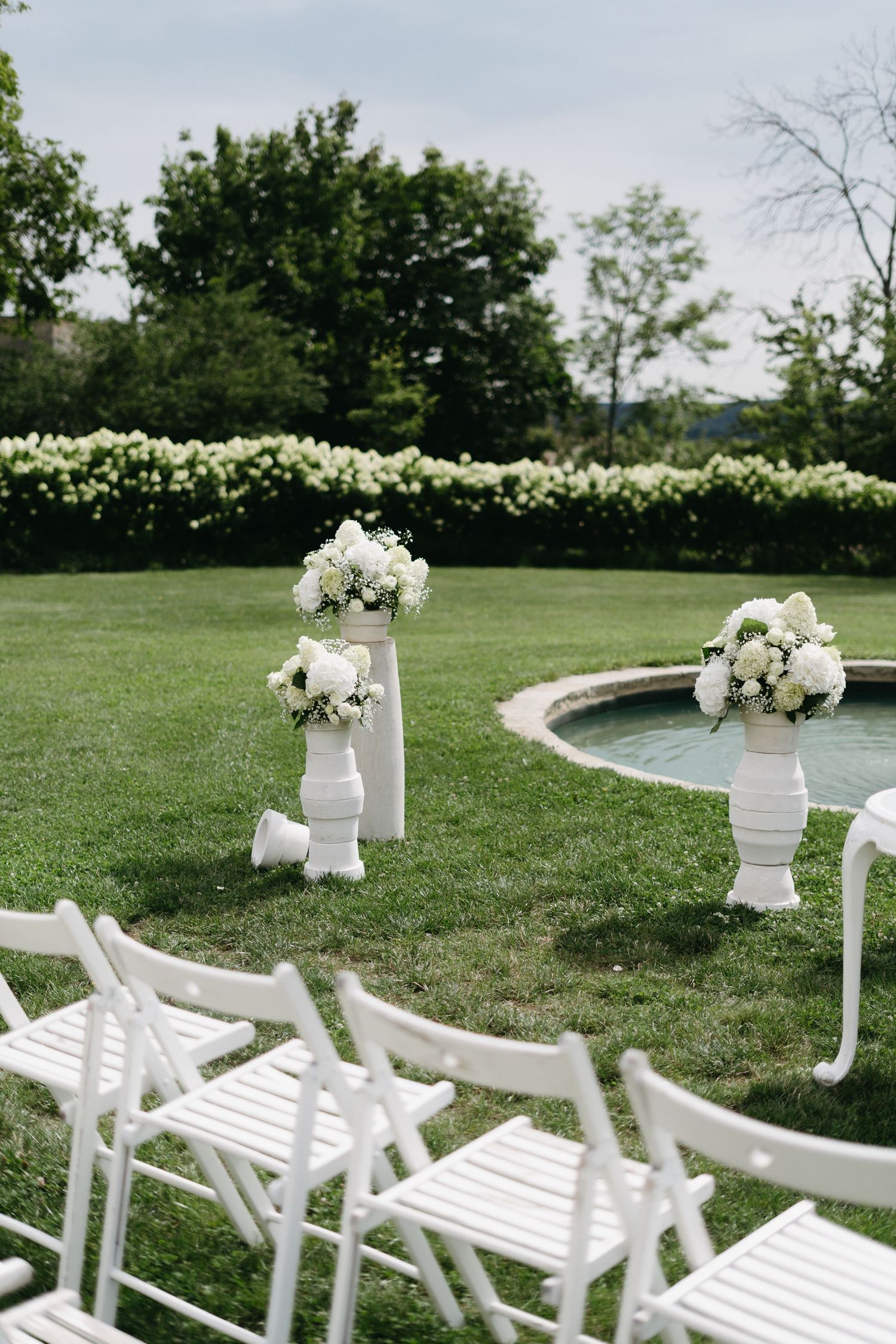 White garden chairs arranged in rows on a lush green lawn with floral decorations for an outdoor wedding ceremony.
