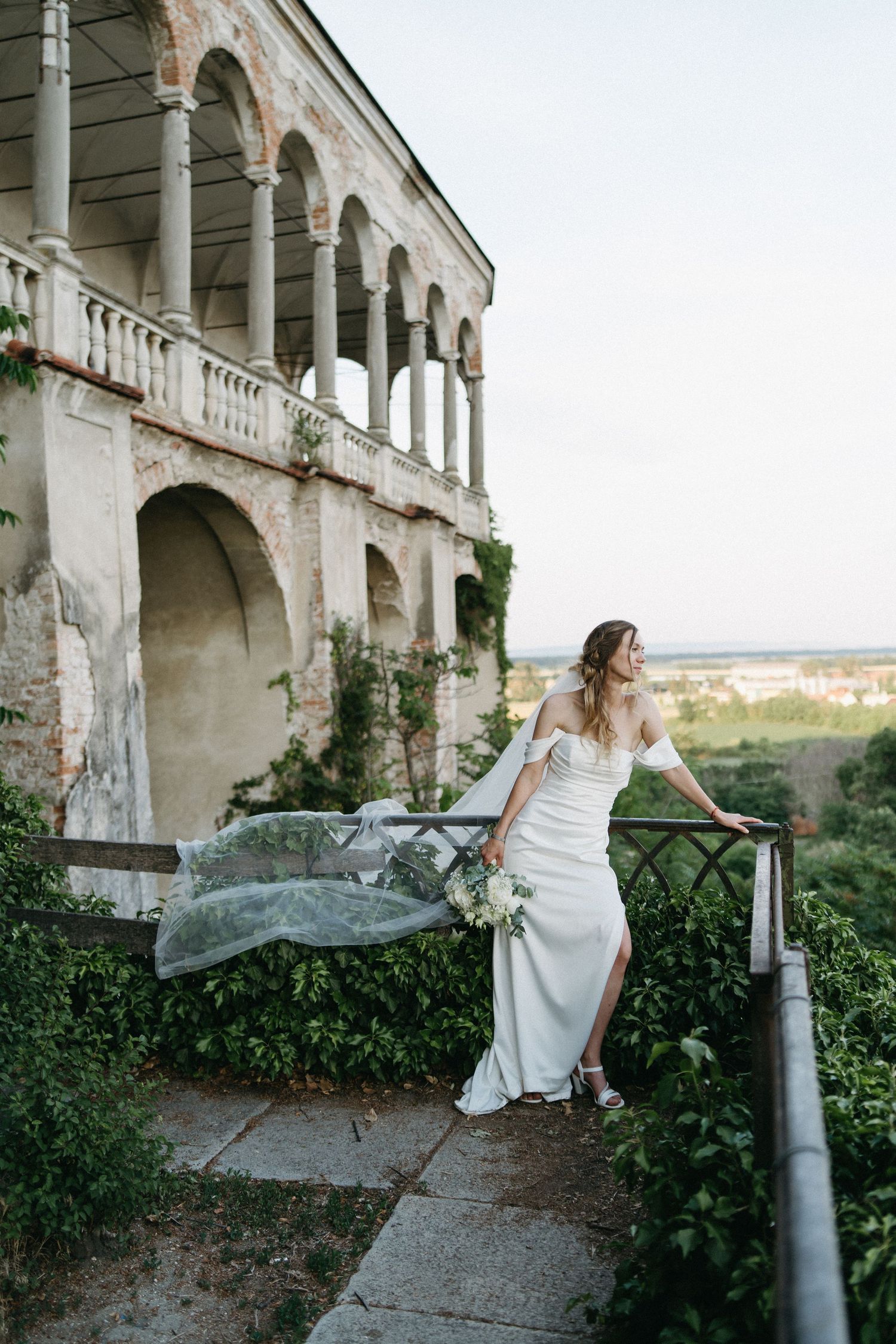 Bride in white off-shoulder gown with flowing veil stands on terrace of historic villa surrounded by greenery.