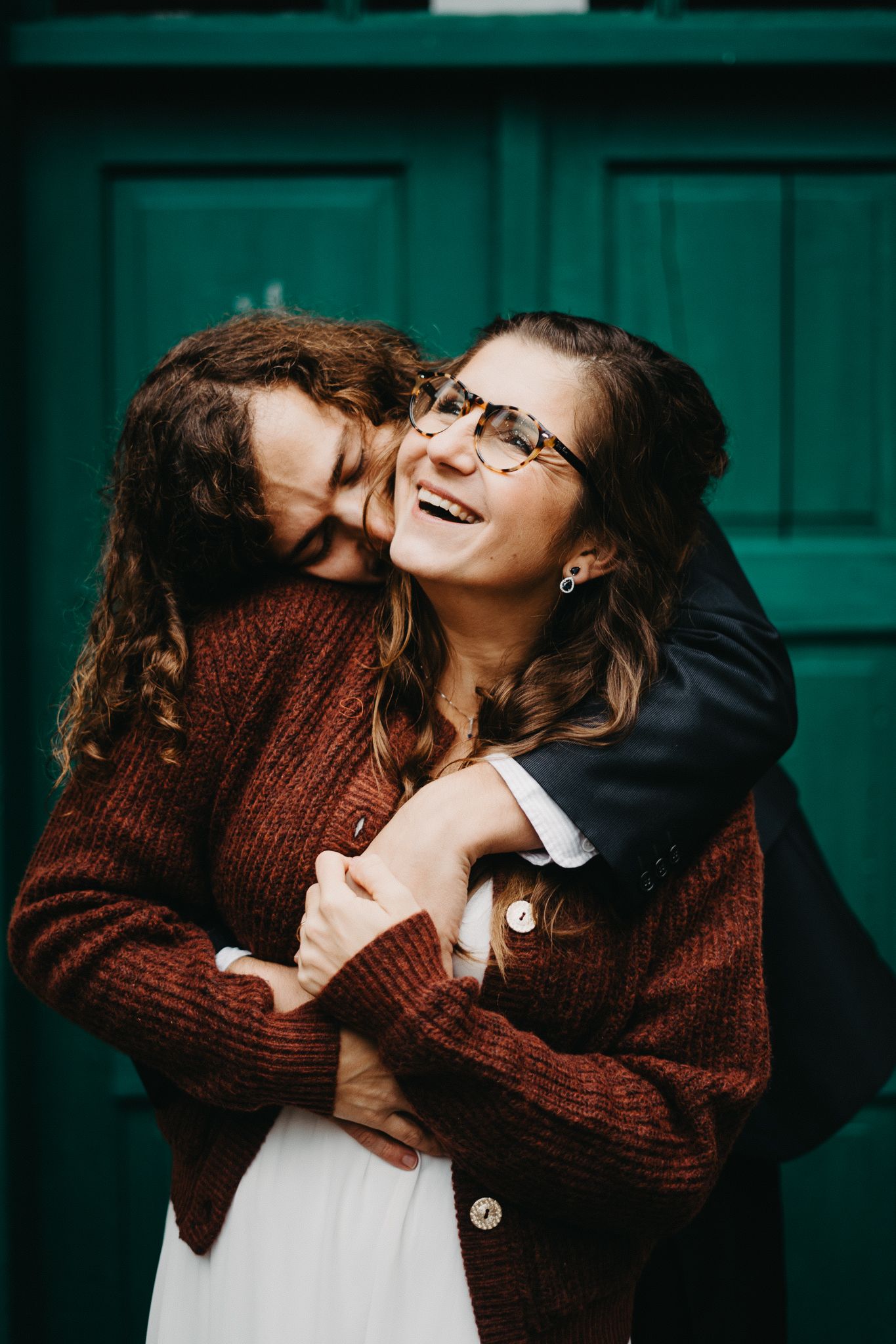 A couple shares a joyful embrace while wearing cozy winter sweaters against a teal-colored background.