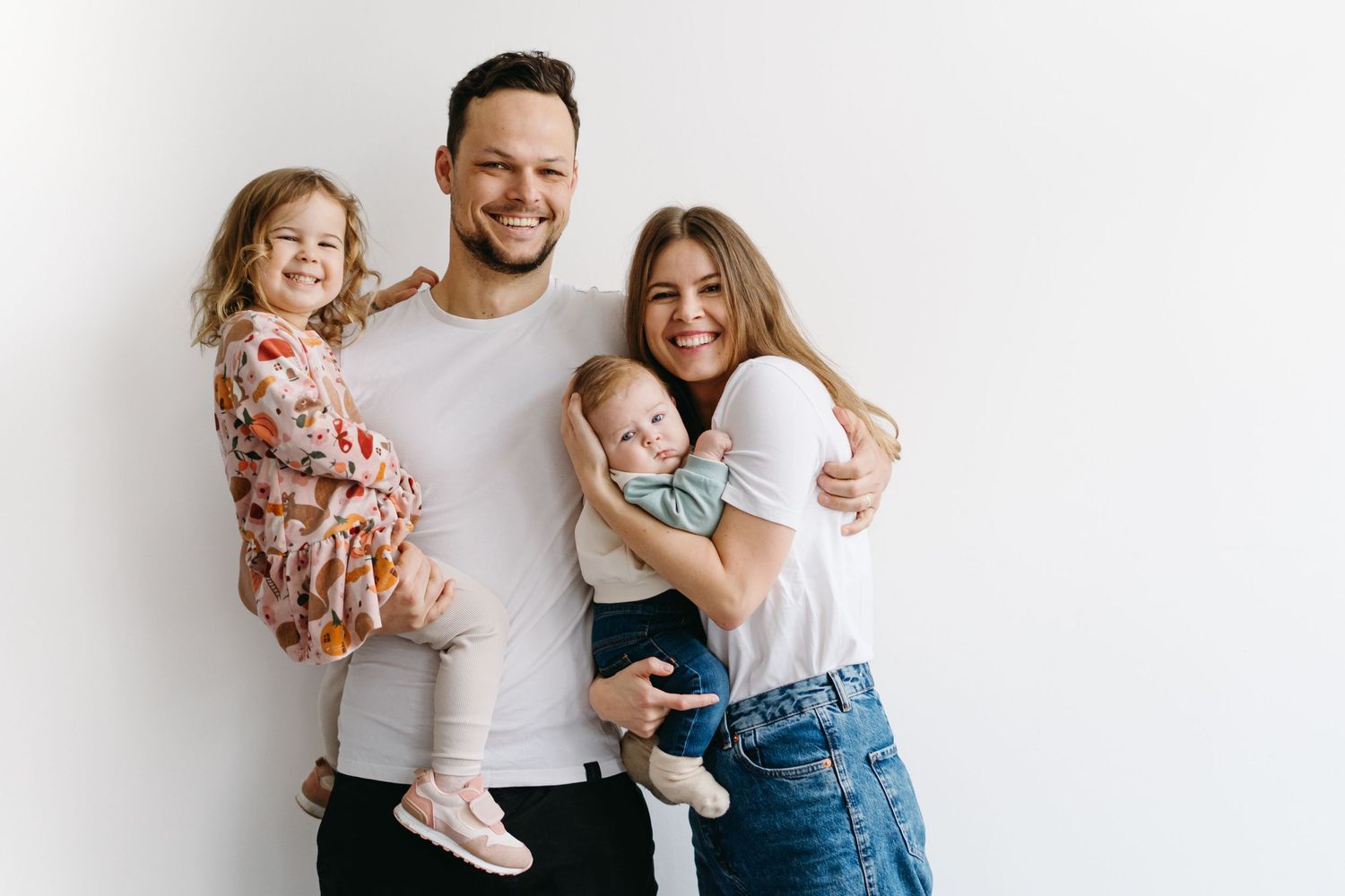 A family of four poses together against a white wall in casual attire.
