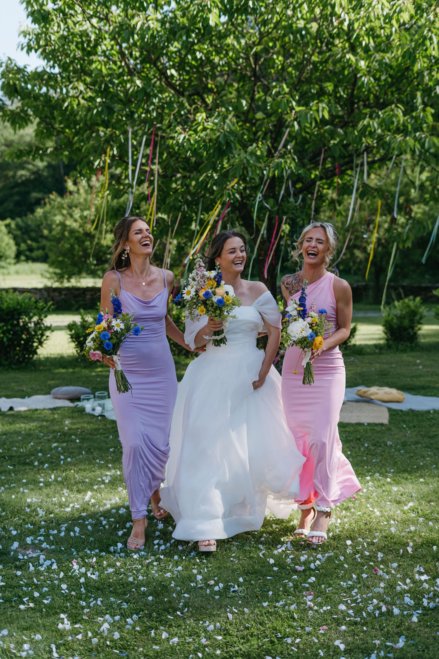 Three women in purple and pink bridesmaid dresses and white wedding gown walk together holding colorful bouquets in a garden.