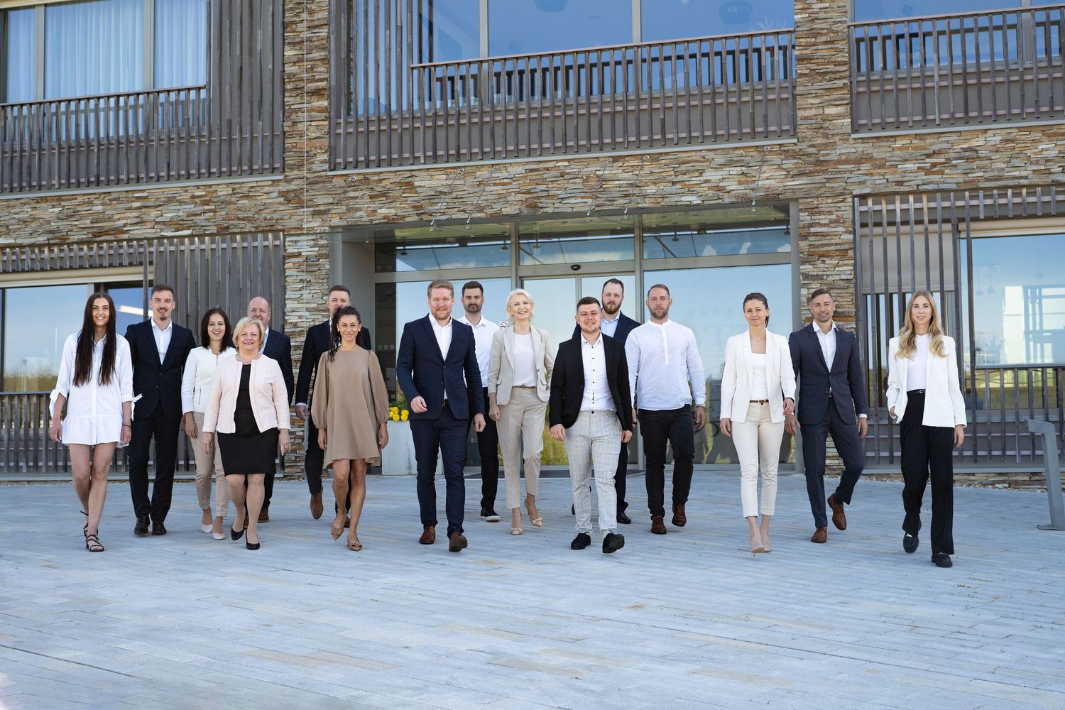 A group of business professionals in formal attire standing together outside a modern building with columns and railings.