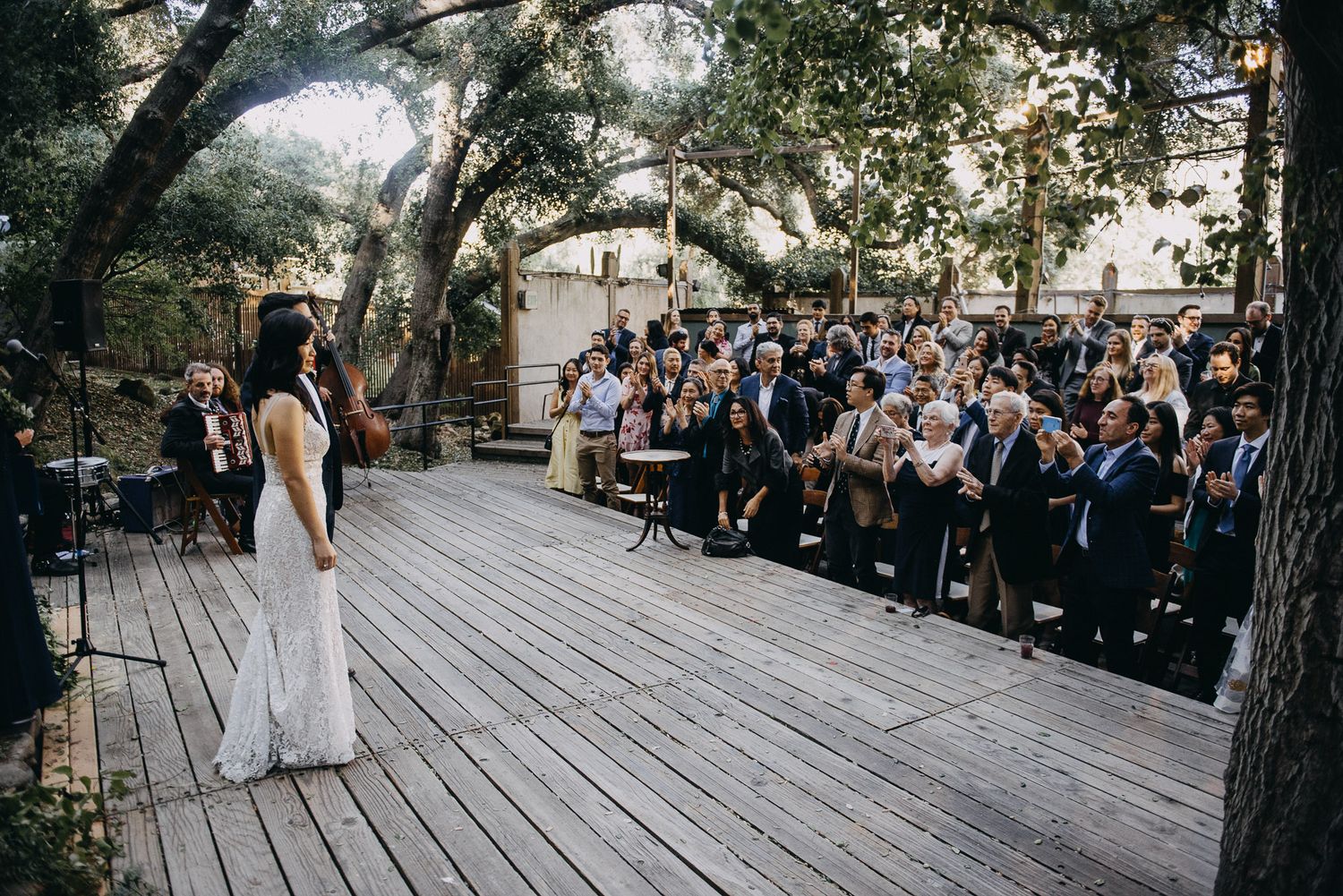 Wedding ceremony guests seated on wooden benches under trees at an outdoor venue.