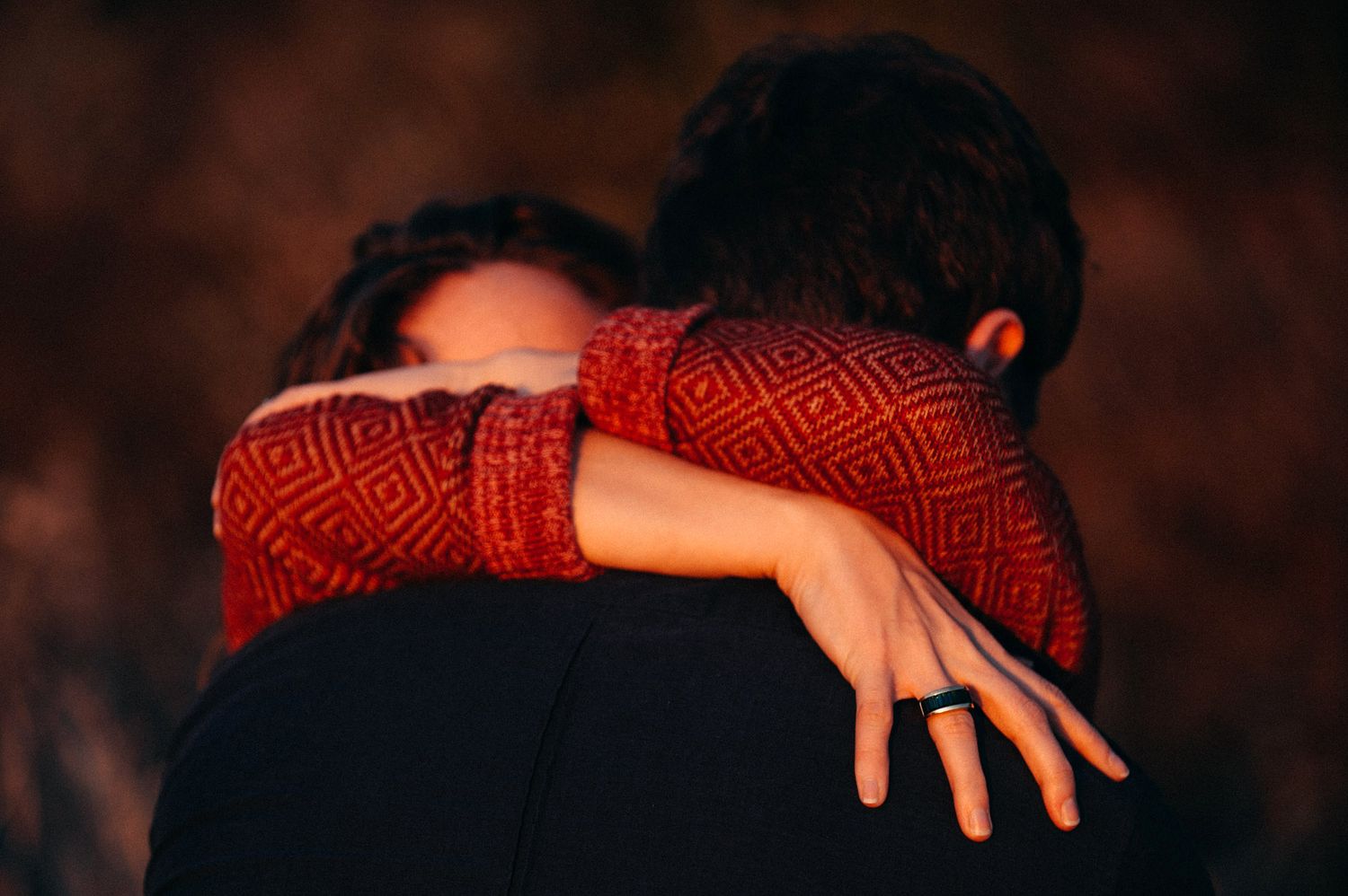 Two people embrace in an intimate moment wearing red patterned clothing against a dark background.