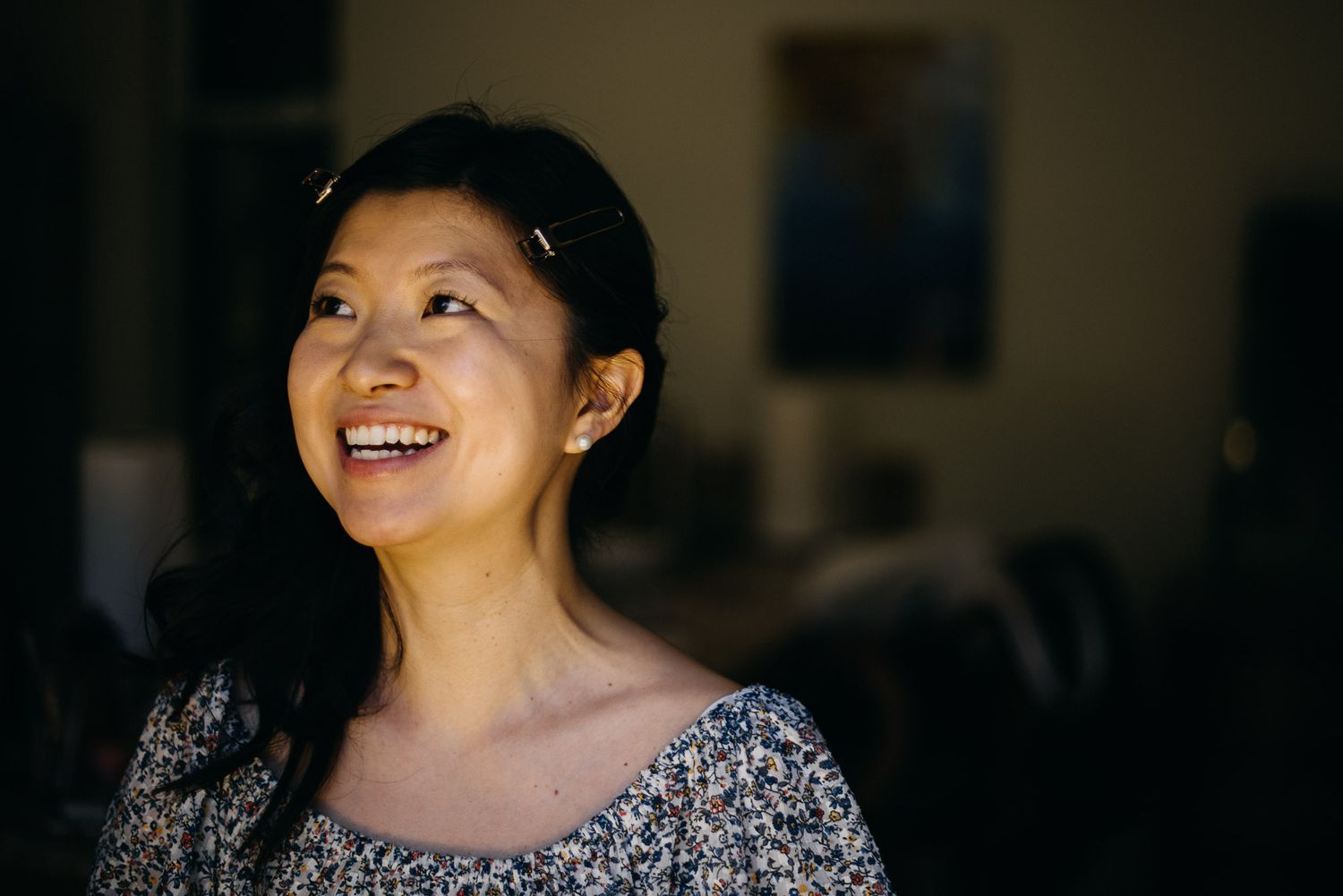 A person in a sparkly top smiles brightly while looking up, illuminated by natural lighting in a dark room.