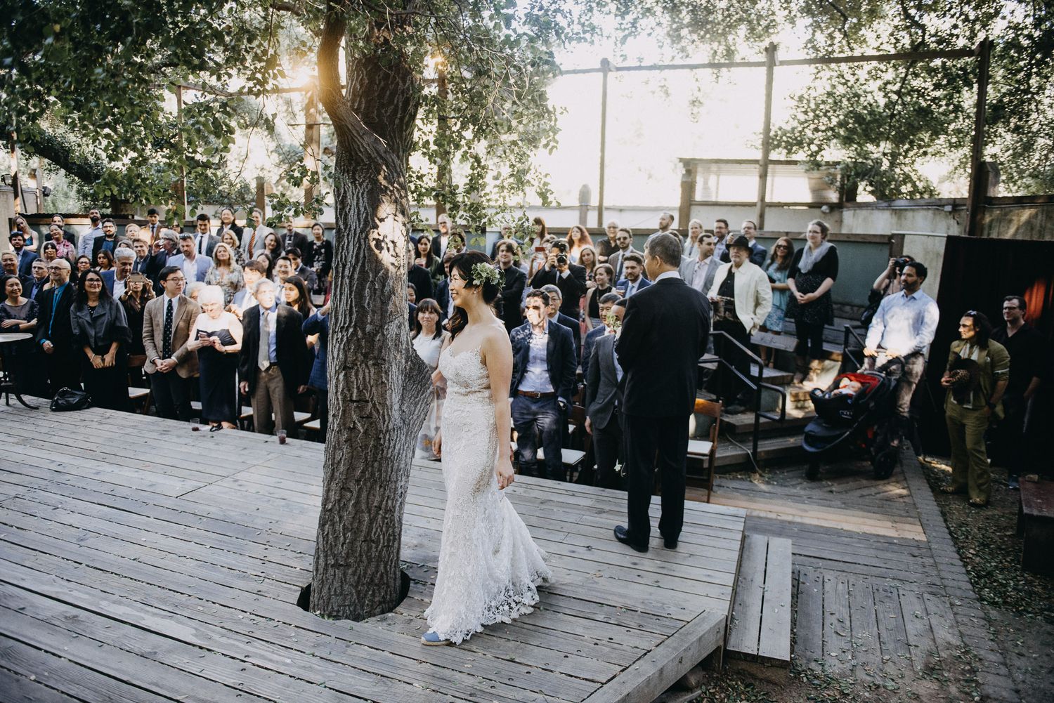 A wedding ceremony takes place on a rustic wooden deck surrounded by trees and guests in formal attire.