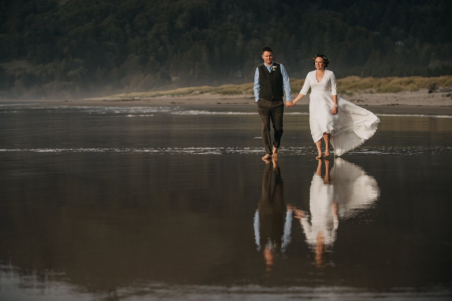 A couple walking on a misty beach at sunset with their reflection mirrored in the wet sand.