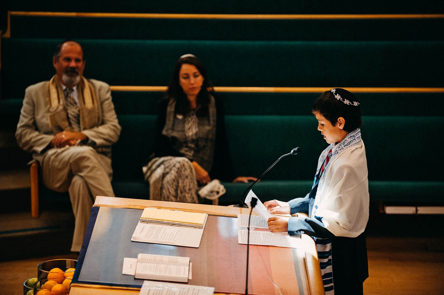 Young person at podium speaking to audience in an auditorium with others seated on green bench.