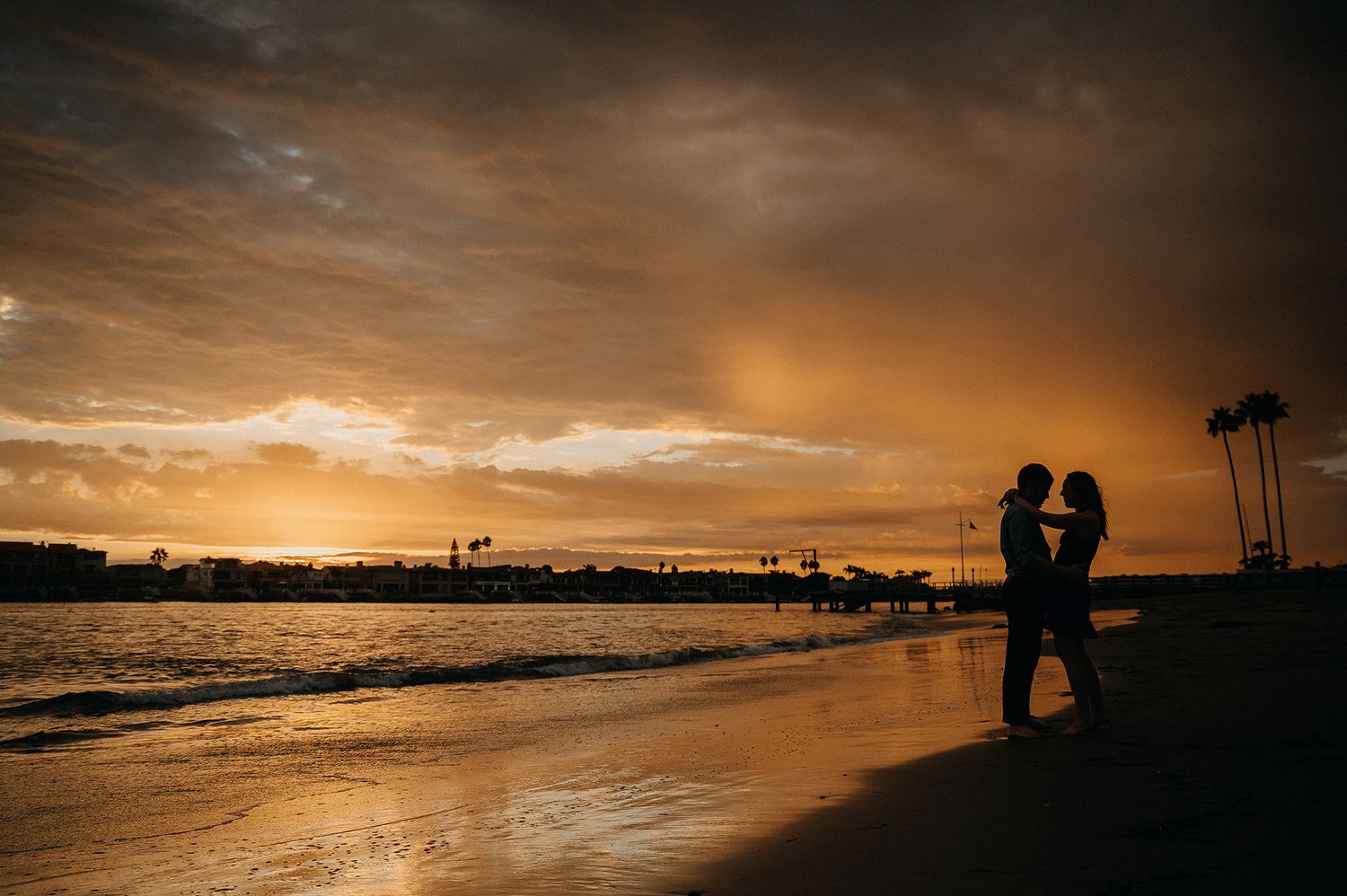 Silhouetted couple stands embracing on a beach at sunset with golden clouds and palm trees reflected in wet sand.