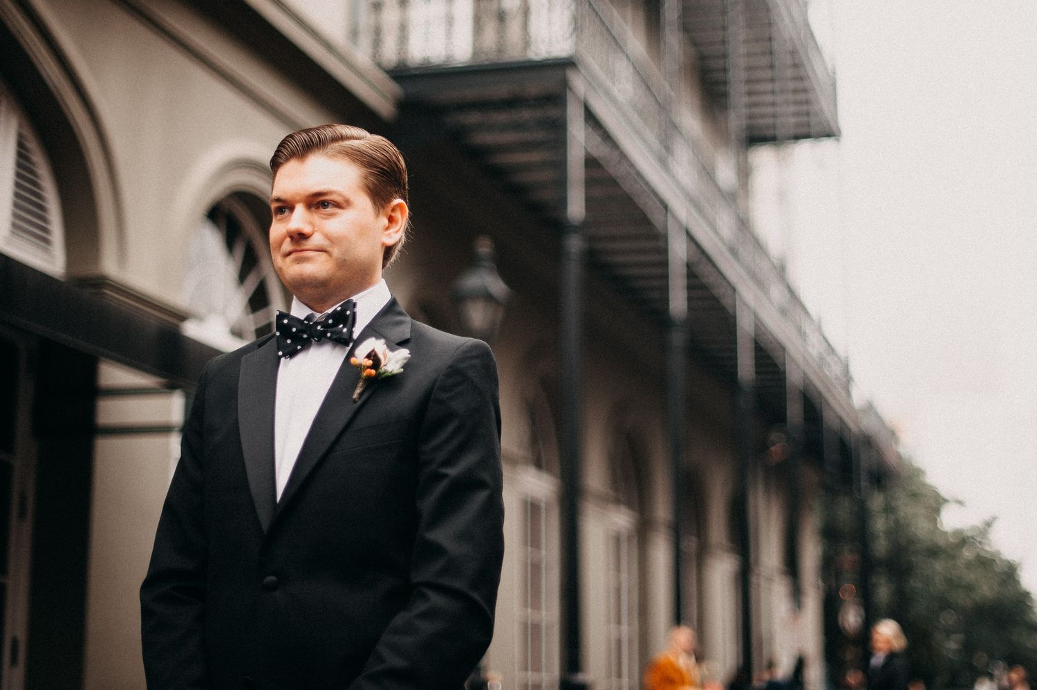 A groom in a classic black tuxedo and bowtie poses against a historic building facade with elegant architecture.