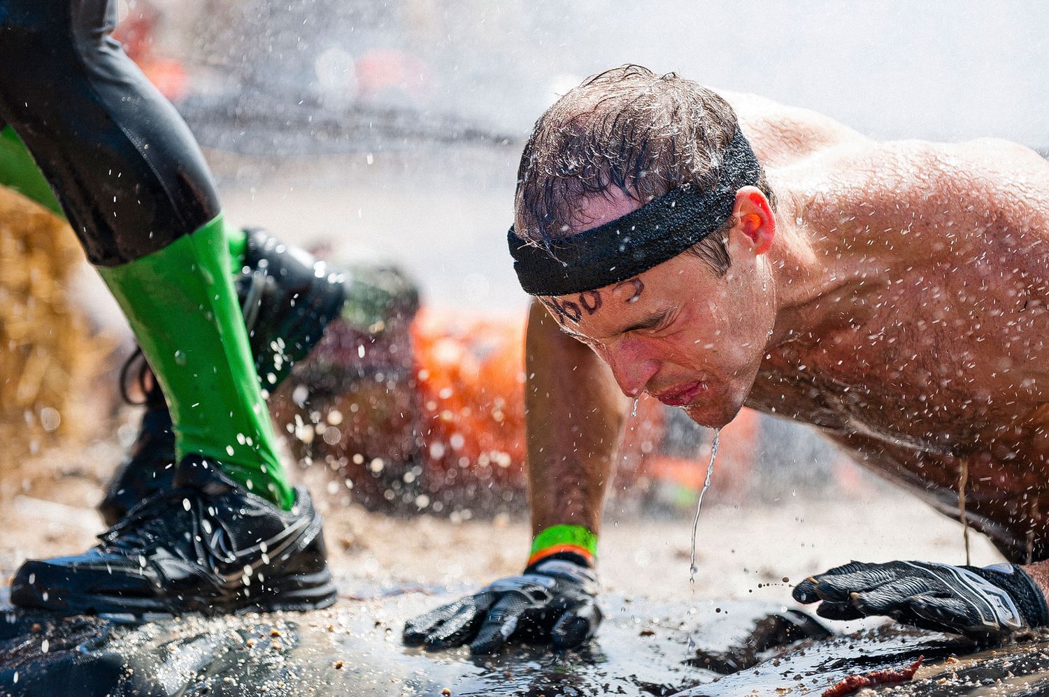 A participant crawls through muddy water during an intense obstacle course race wearing neon green and black gear.