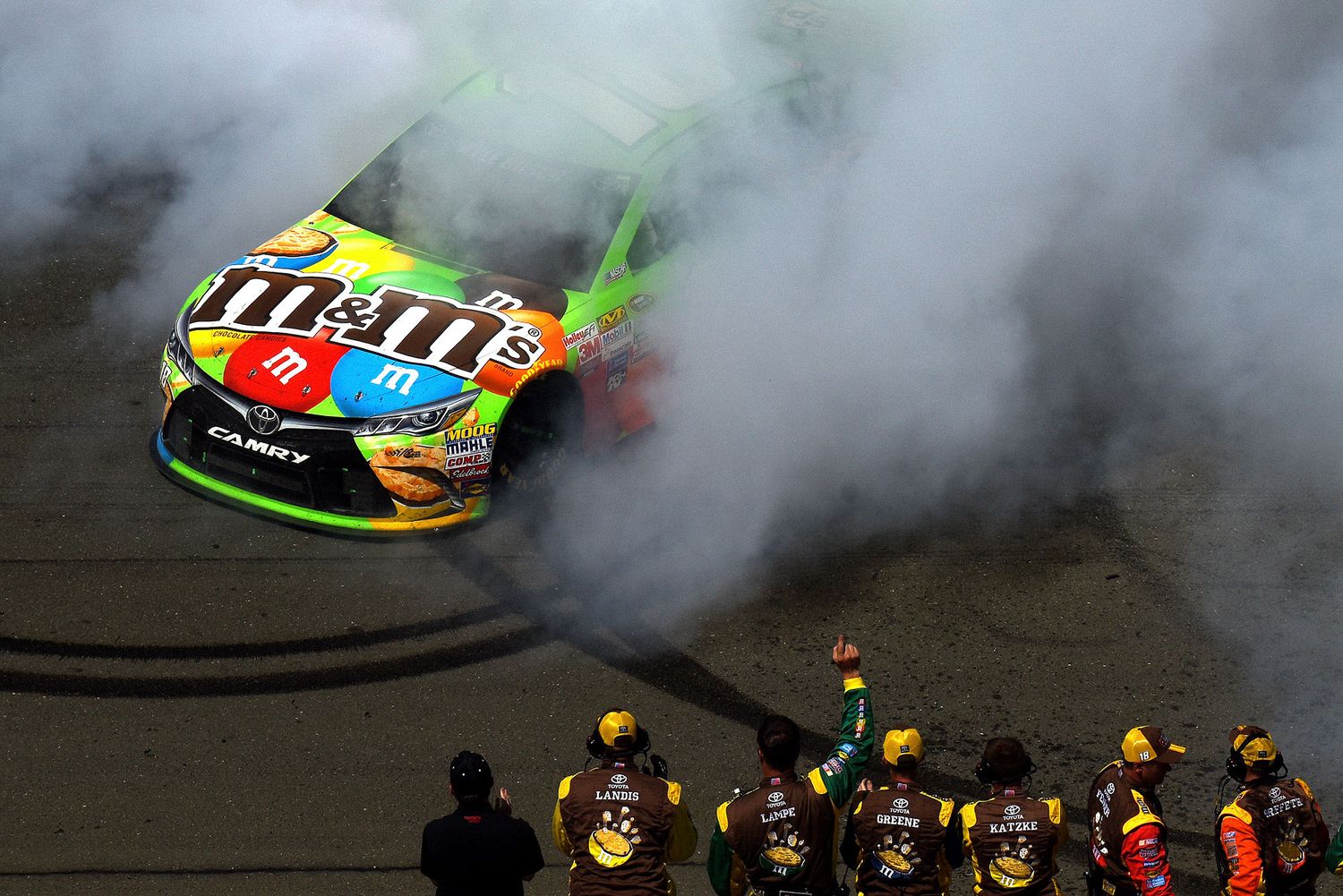 M&M's NASCAR race car creates smoke while celebrating victory with pit crew watching.
