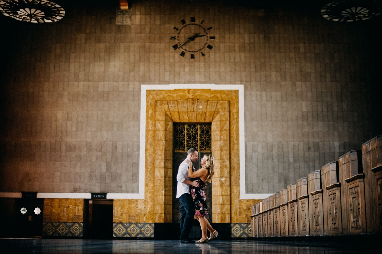 A romantic couple embraces in front of an ornate yellow doorway with vintage lockers in the background.