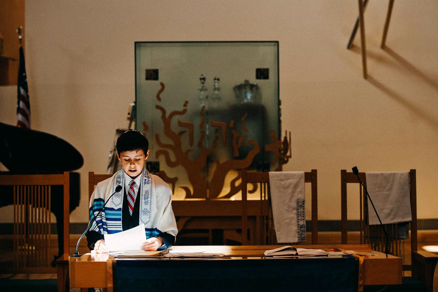 A person in professional attire sits at a desk reading documents in a dimly lit room with art displayed on the wall.