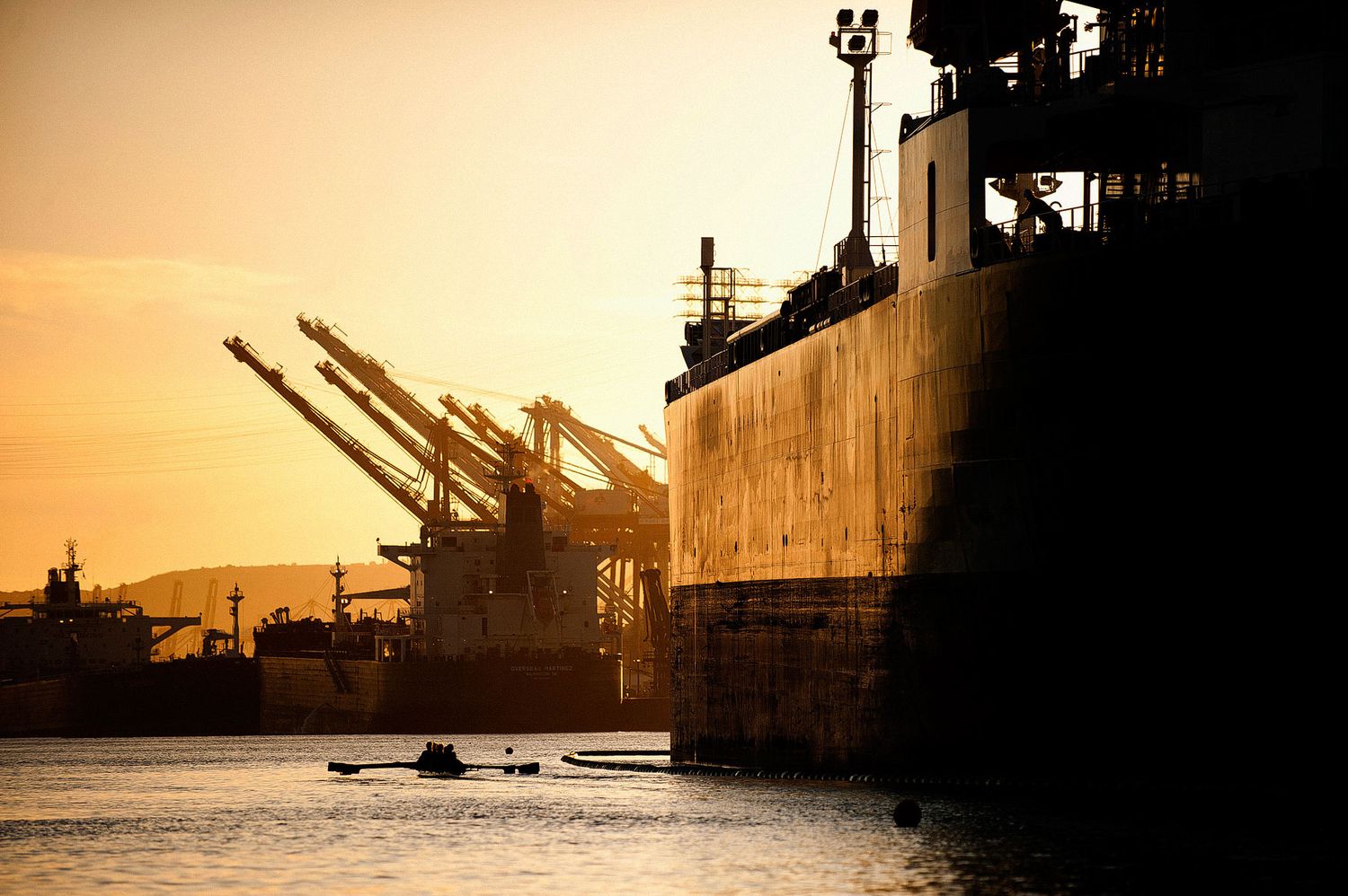 Silhouette of cargo ship with cranes at port during golden sunset with water in foreground.