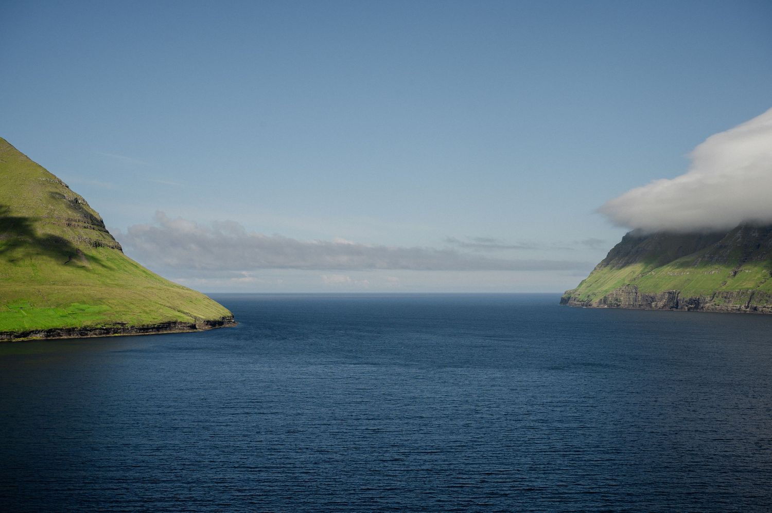 Deep blue waters flow between steep green coastal cliffs under a bright sky in the Faroe Islands.