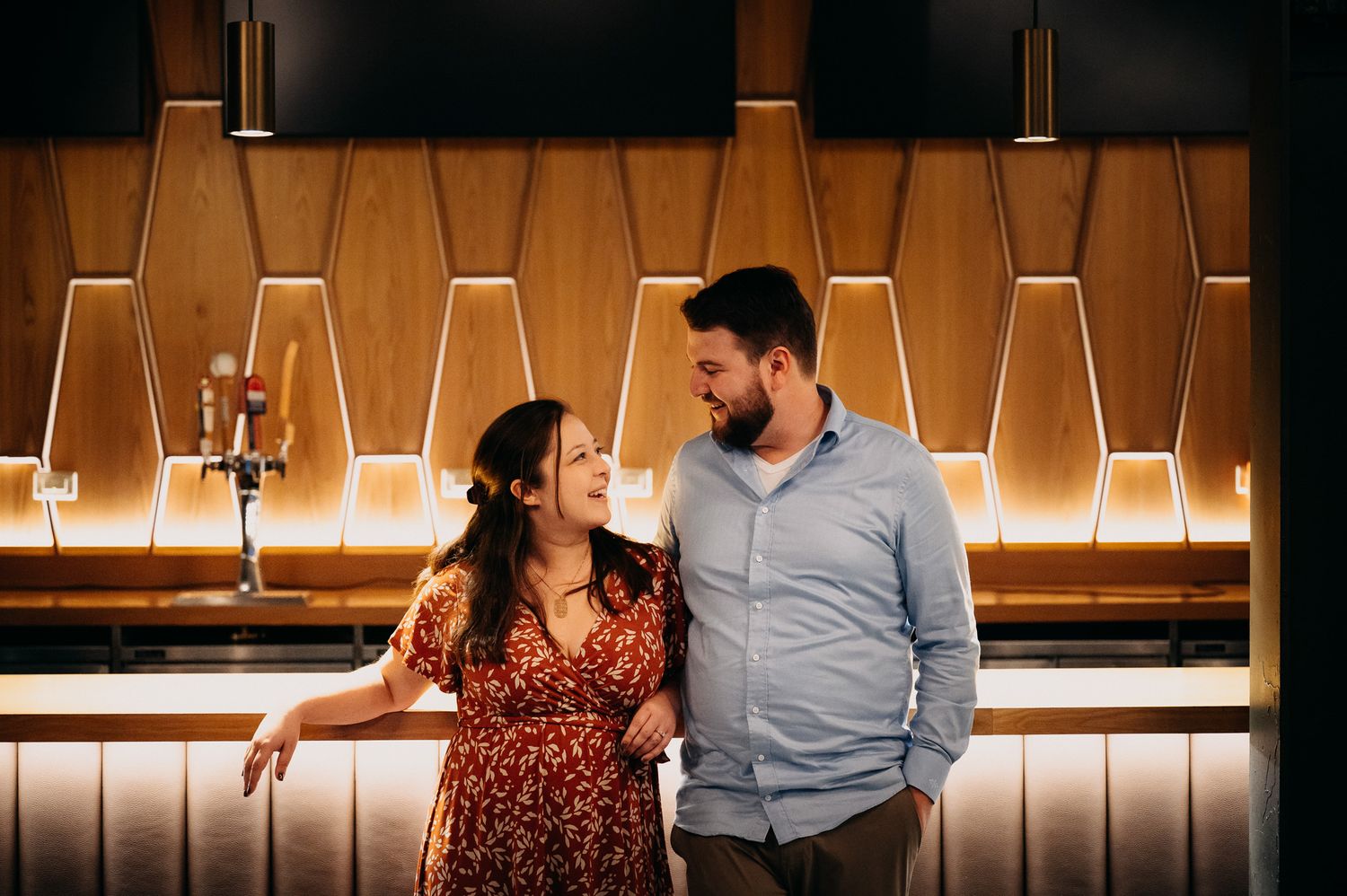 Two people stand together at a modern bar with illuminated geometric wooden panels on the wall behind them.