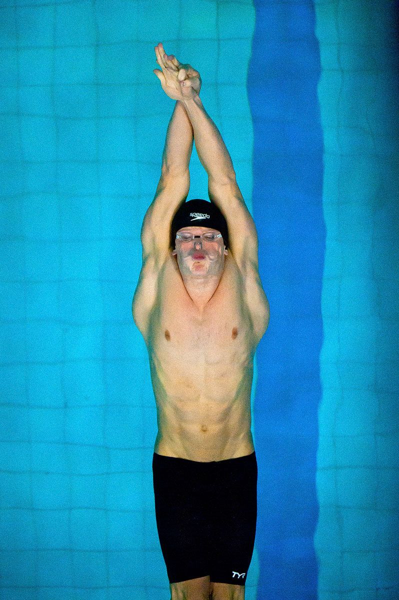 A competitive swimmer in black swimwear stretches upward against a bright turquoise pool background.