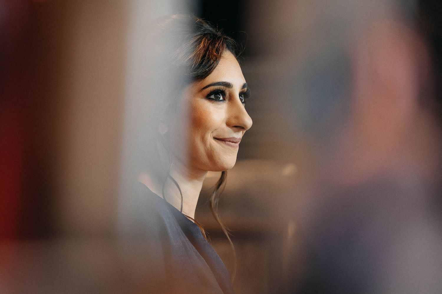 Side profile portrait of someone with dark hair smiling while listening with earbuds in a soft-focus setting.