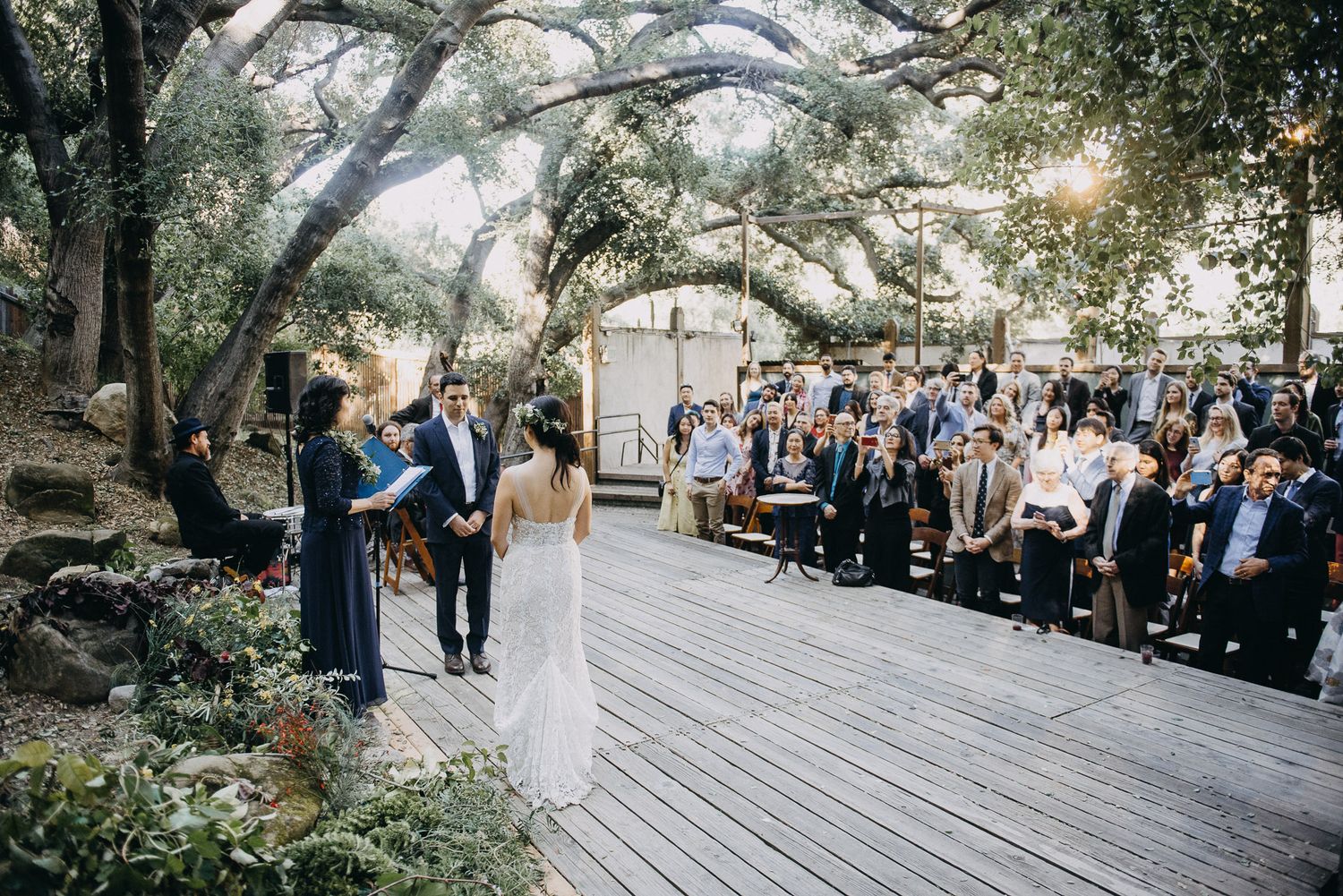 An outdoor wedding ceremony takes place on a wooden deck surrounded by trees with guests seated on benches.