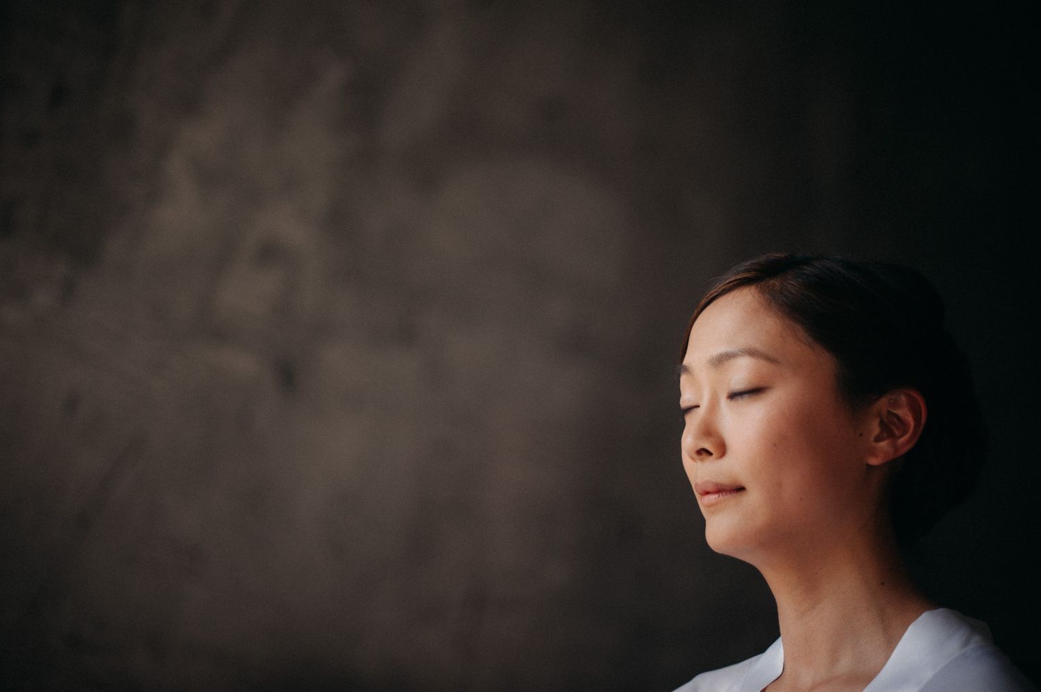 Person with closed eyes meditating against dark textured wall in peaceful contemplative pose.