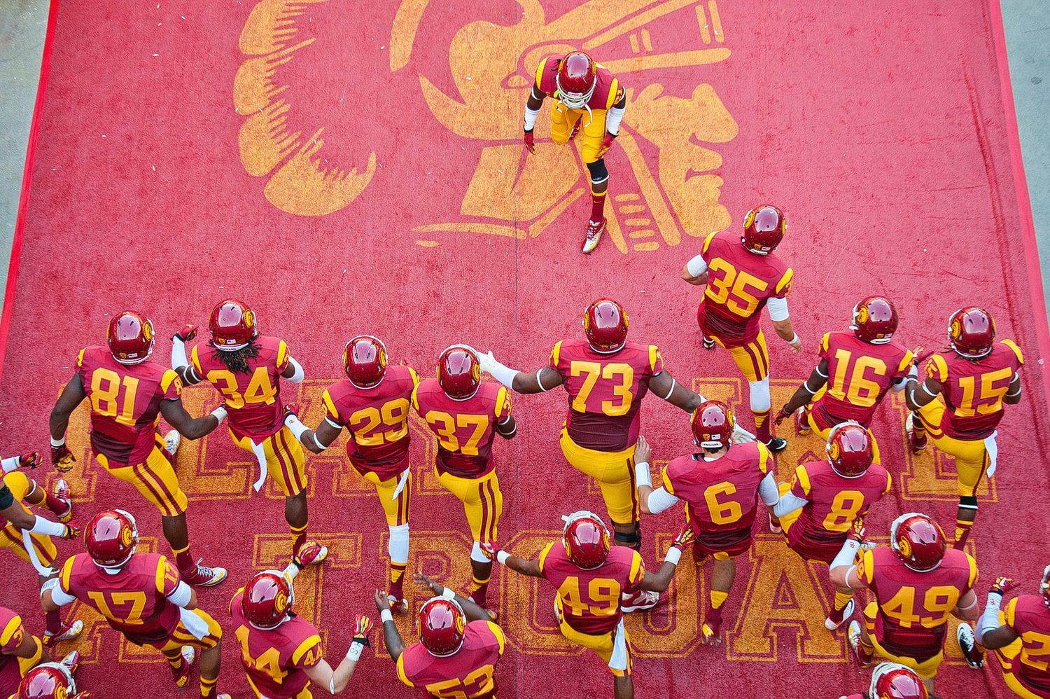 Aerial view of USC football team in cardinal and gold uniforms gathered on pink field.