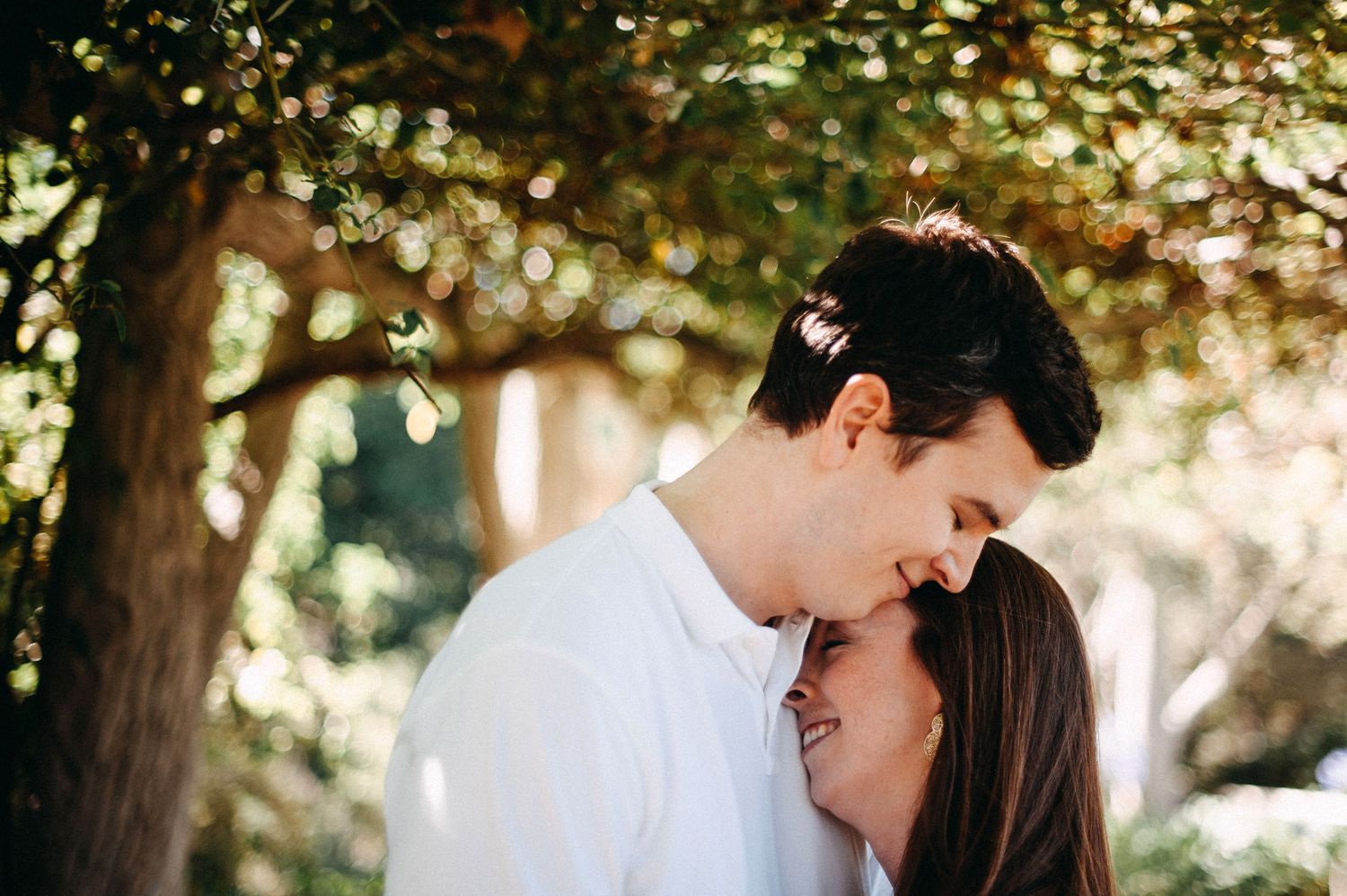 Couple shares tender moment in dappled sunlight beneath trees in romantic outdoor setting.