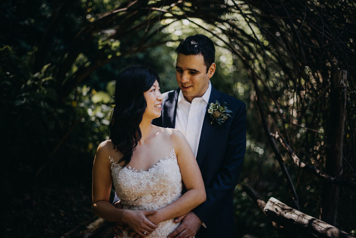 Couple in wedding attire share an intimate moment in a forest setting with dappled sunlight.