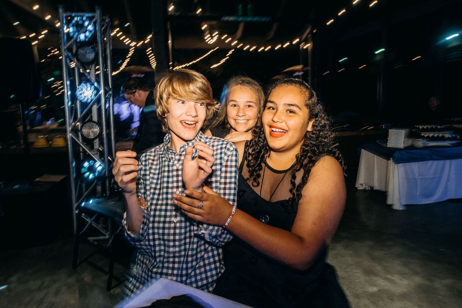 Group of friends smiling and taking selfies at an evening party with string lights in background.