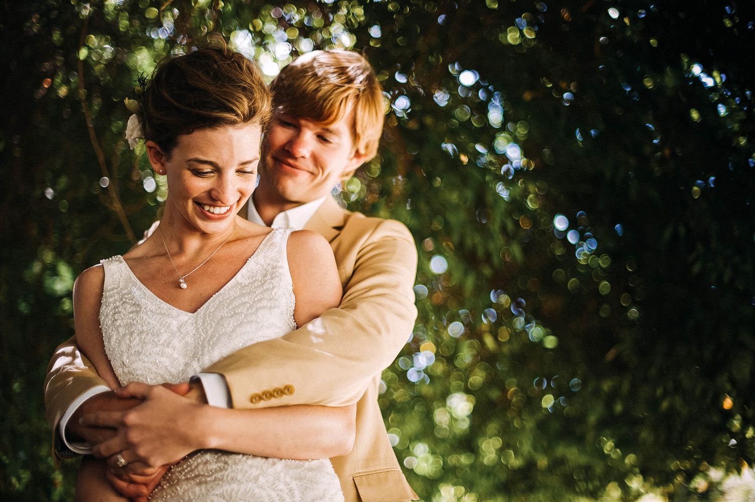 A couple embraces outdoors under dappled sunlight and trees, with the person in white wearing a lace dress.