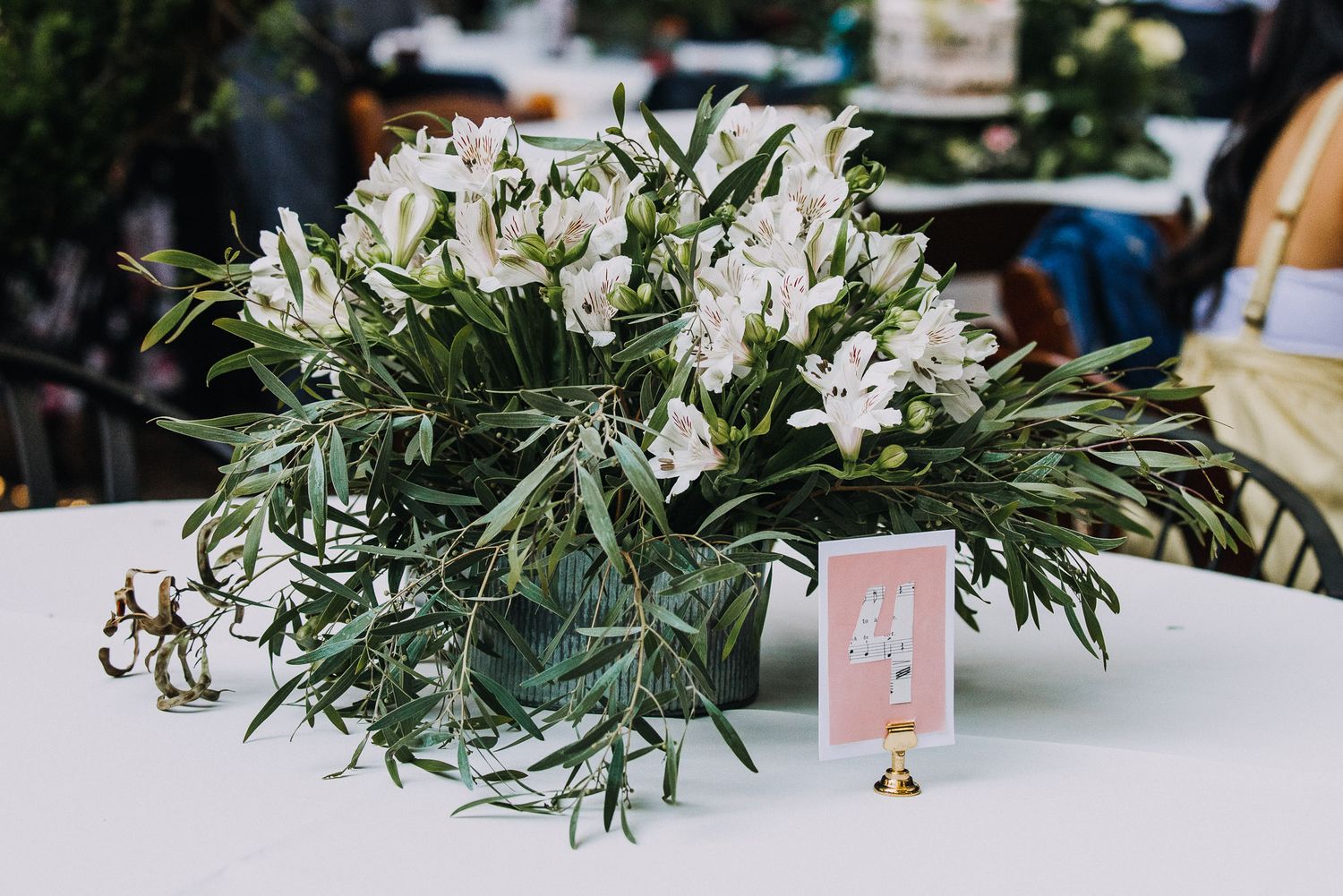 White floral centerpiece with greenery and table number card displayed on white tablecloth at event setting.