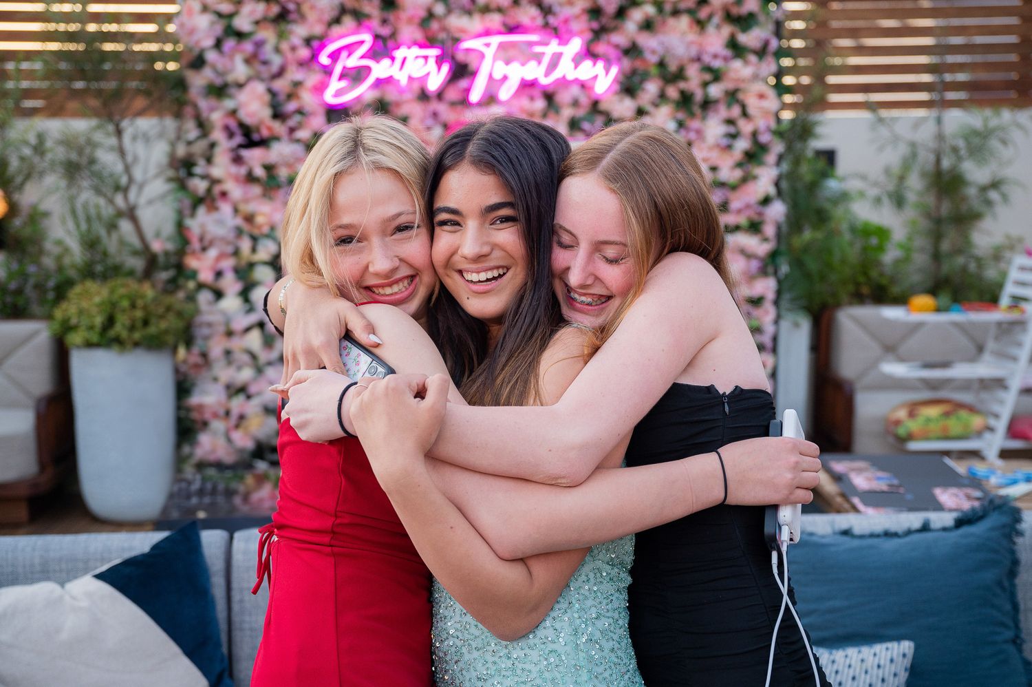 Three friends share a joyful embrace while smiling at an outdoor party with pink floral decorations.