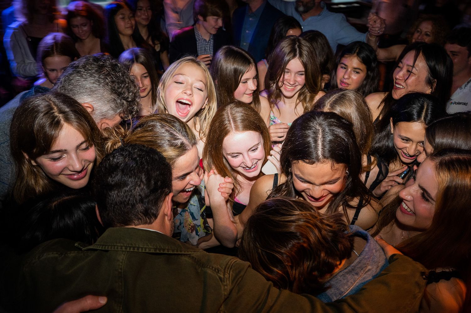 A crowd of people laugh and huddle together at a dimly lit nightclub or party venue.