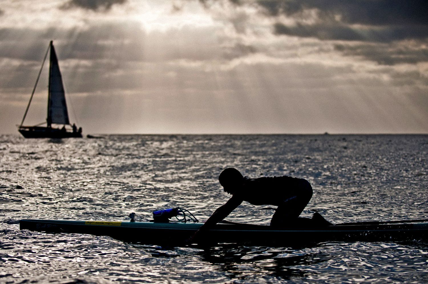 Silhouette of kayaker at sunset with sailboat on horizon and dramatic cloudy sky over ocean.