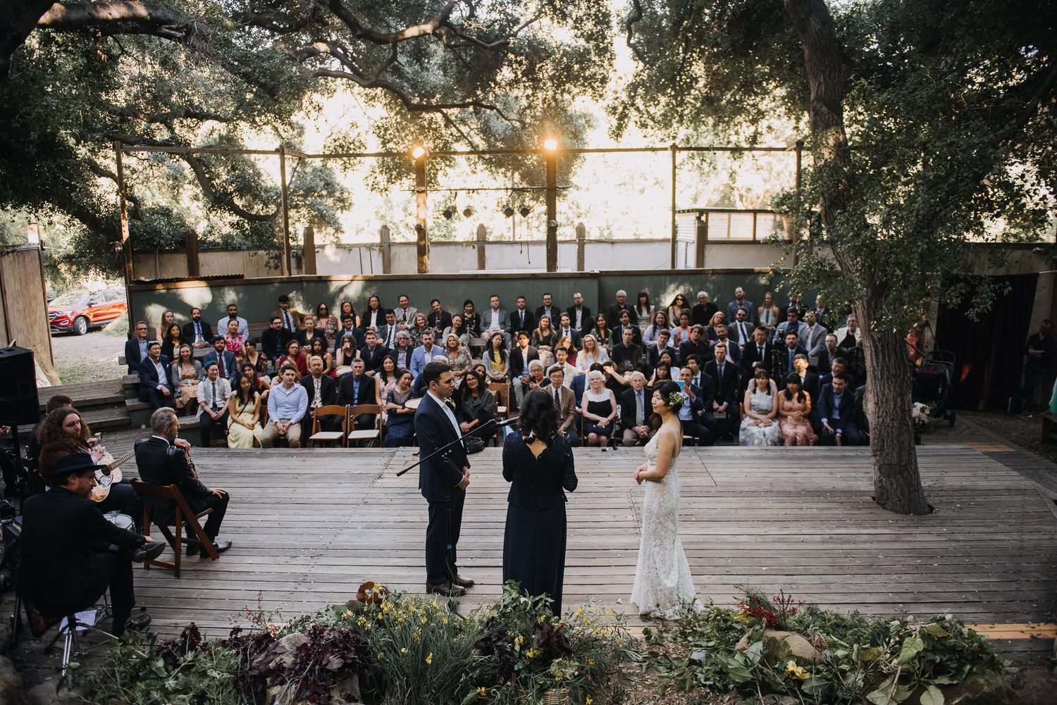 Wedding guests gather outdoors at a scenic venue during a golden hour celebration under tall trees.